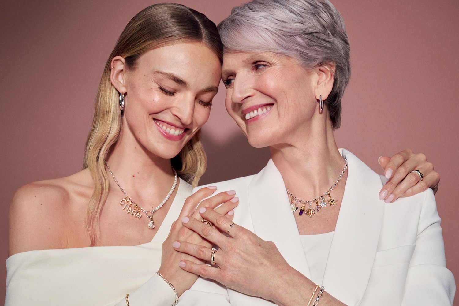 Two women, one younger and one older, smiling and embracing each other, wearing white clothing and jewelry, with a pink background.