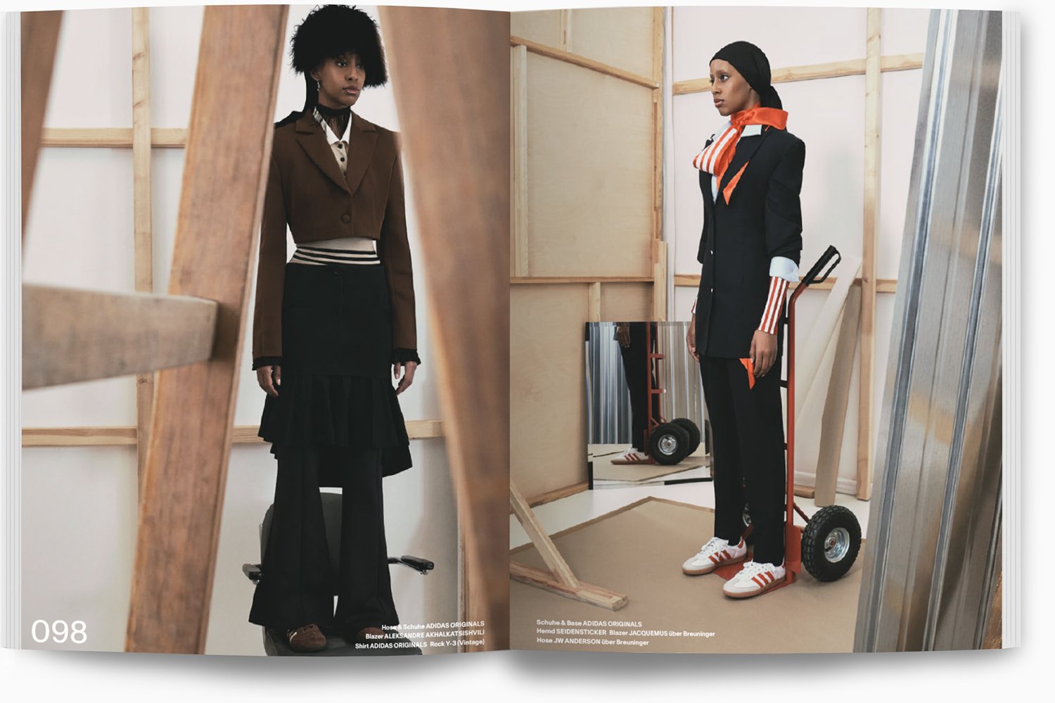 Zwei Frauen in Modeoutfits stehen in einem Raum, der wie eine Werkstatt aussieht, umgeben von Holzrahmen, Spiegeln und Baumaterialien.