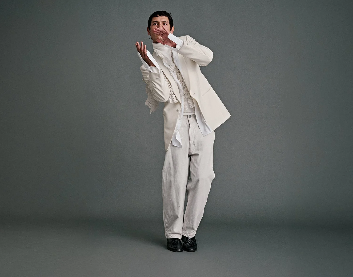 A young man in a white suit covering his mouth and nose with his hand in a pose against a plain gray background.