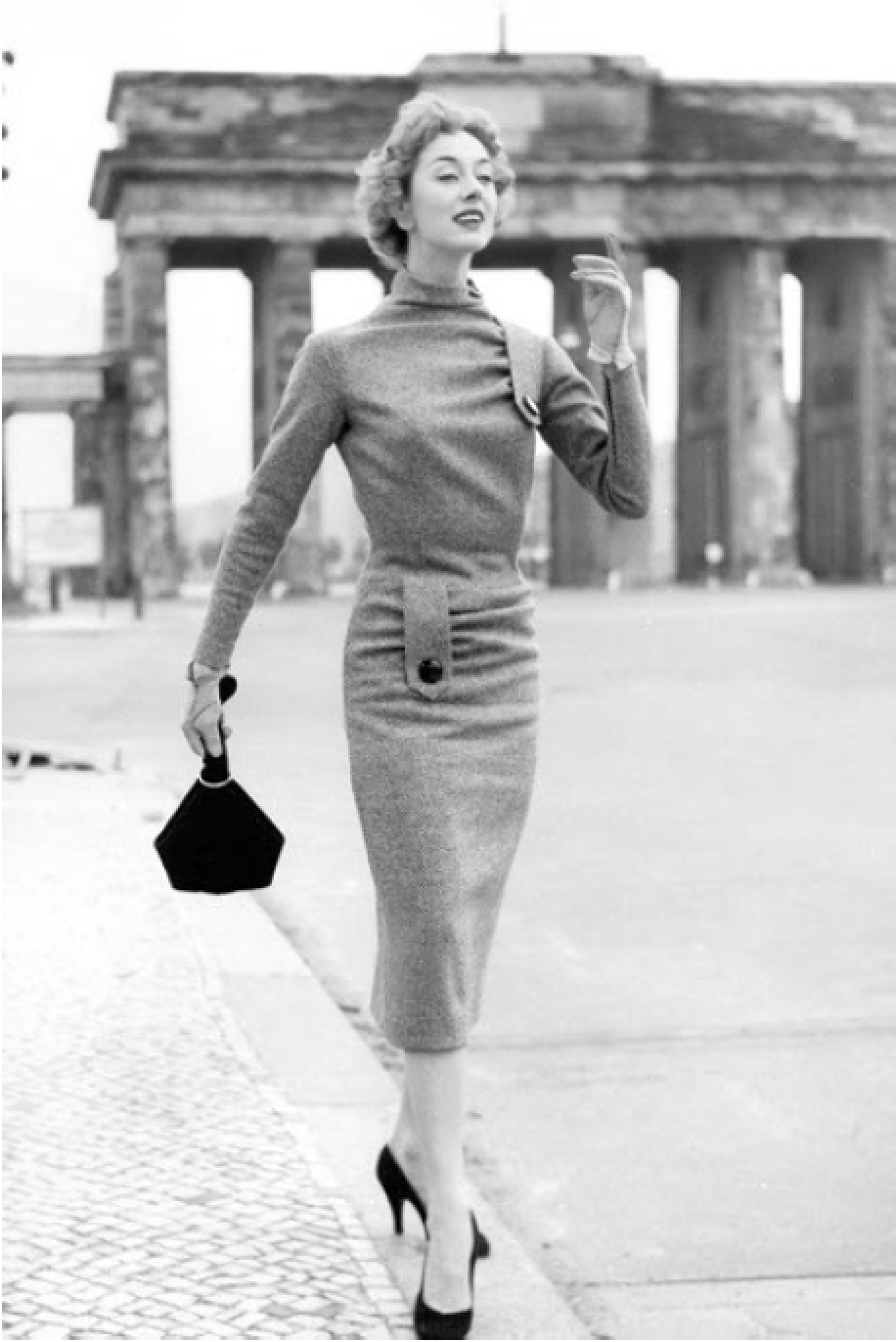 Eine Frau in einem eleganten Kleid geht an einer historischen Gebäudearchitektur in Berlin vorbei, vermutlich dem Brandenburger Tor, im Jahr 1950.