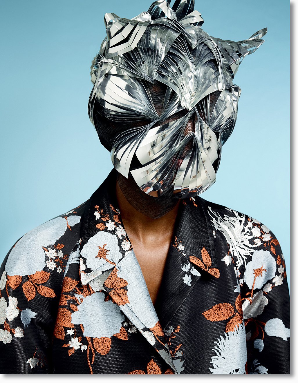 Person trägt eine futuristische, metallische Maske aus reflektierendem Material und einen schwarzen Kimono mit floralen Mustern in Weiß, Hellblau und Orange vor einem hellblauen Hintergrund.