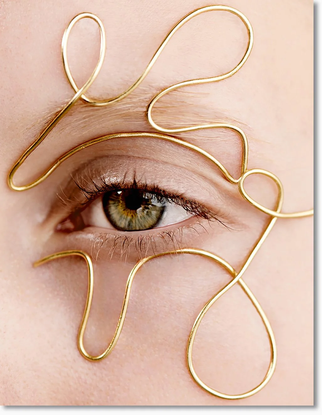 Nahaufnahme eines grauen Auges mit goldfarbenem Draht, der kunstvoll die Form eines Gesichts oder Musters um das Auge bildet.