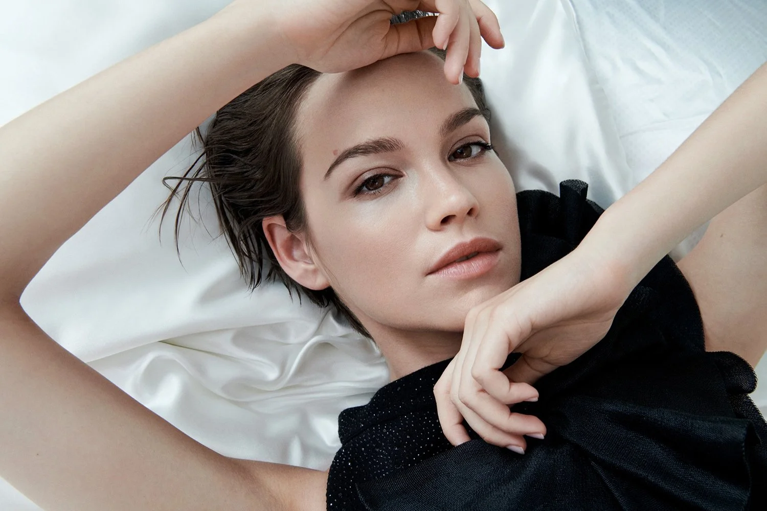 Close-up of a woman with short brown hair lying on a white pillow, wearing a black top, with one hand resting on her head and the other near her face, looking directly at the camera.