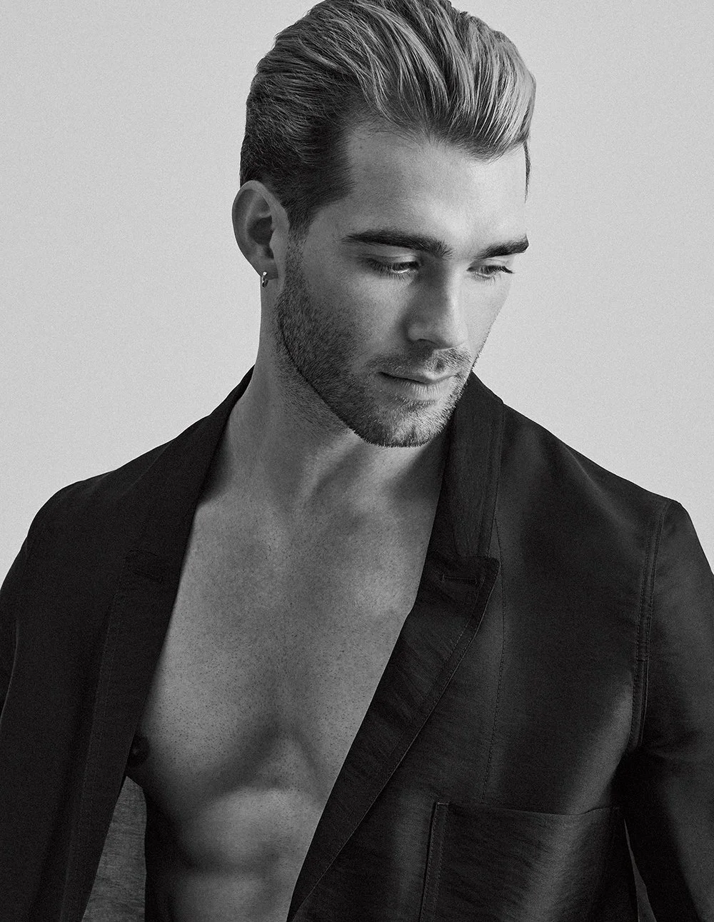 Schwarz-weiß Porträt eines jungen Mannes mit gepflegtem Bart und kurzen, nach hinten gestylten Haaren, trägt ein offenes schwarzes Jackett und sieht nach unten.