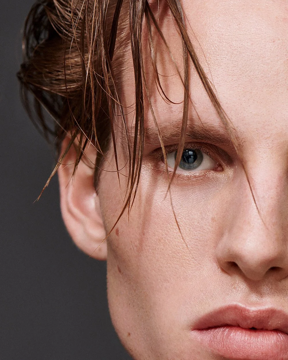 Nahaufnahme eines jungen Mannes mit nassen, braunen Haaren und blauen Augen. Nur die rechte Gesichtshälfte ist sichtbar, vor einem dunklen Hintergrund.