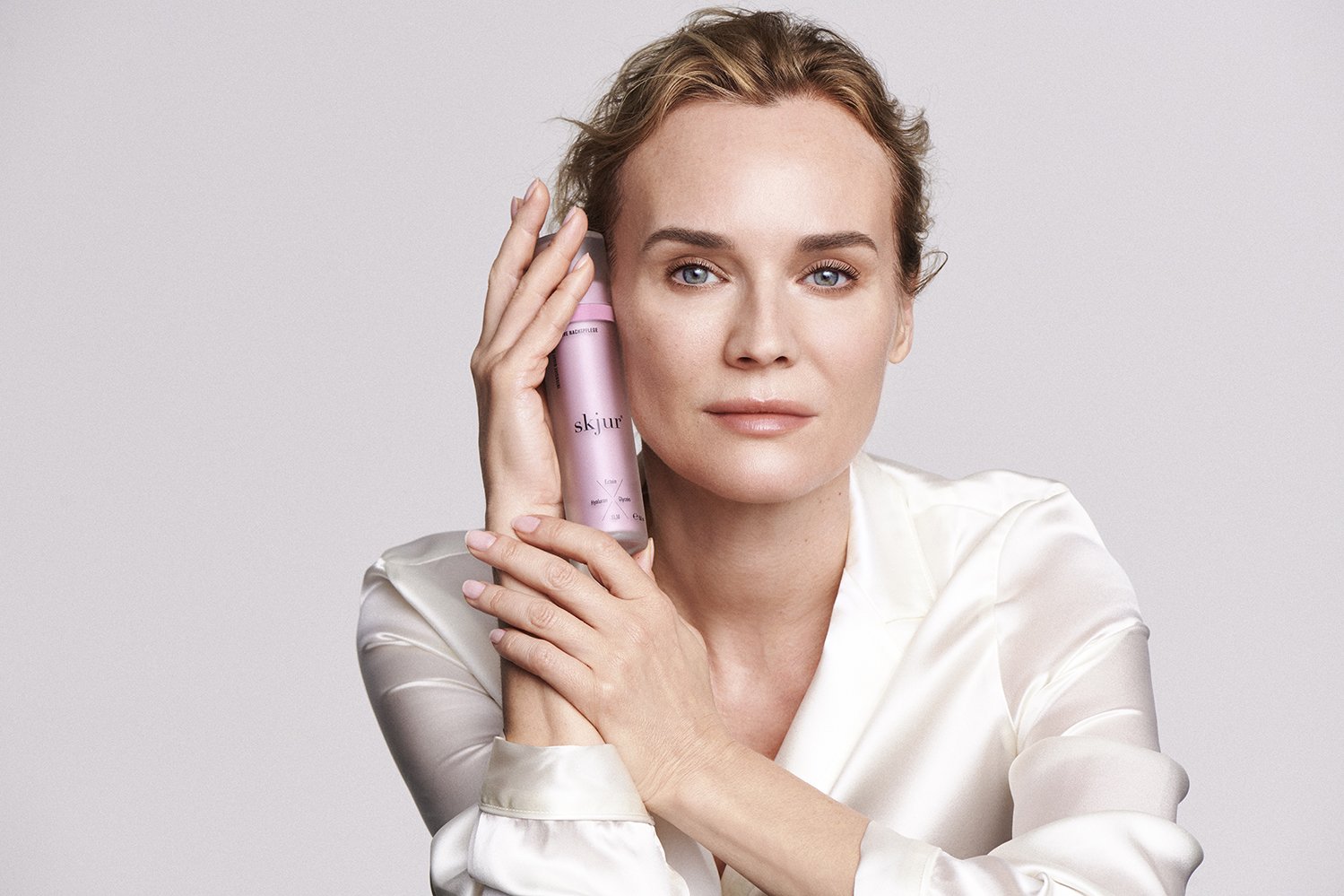 A woman with fair skin and blue eyes holds a pink skincare product bottle to her face, wearing a silky white blouse, against a plain light background.
