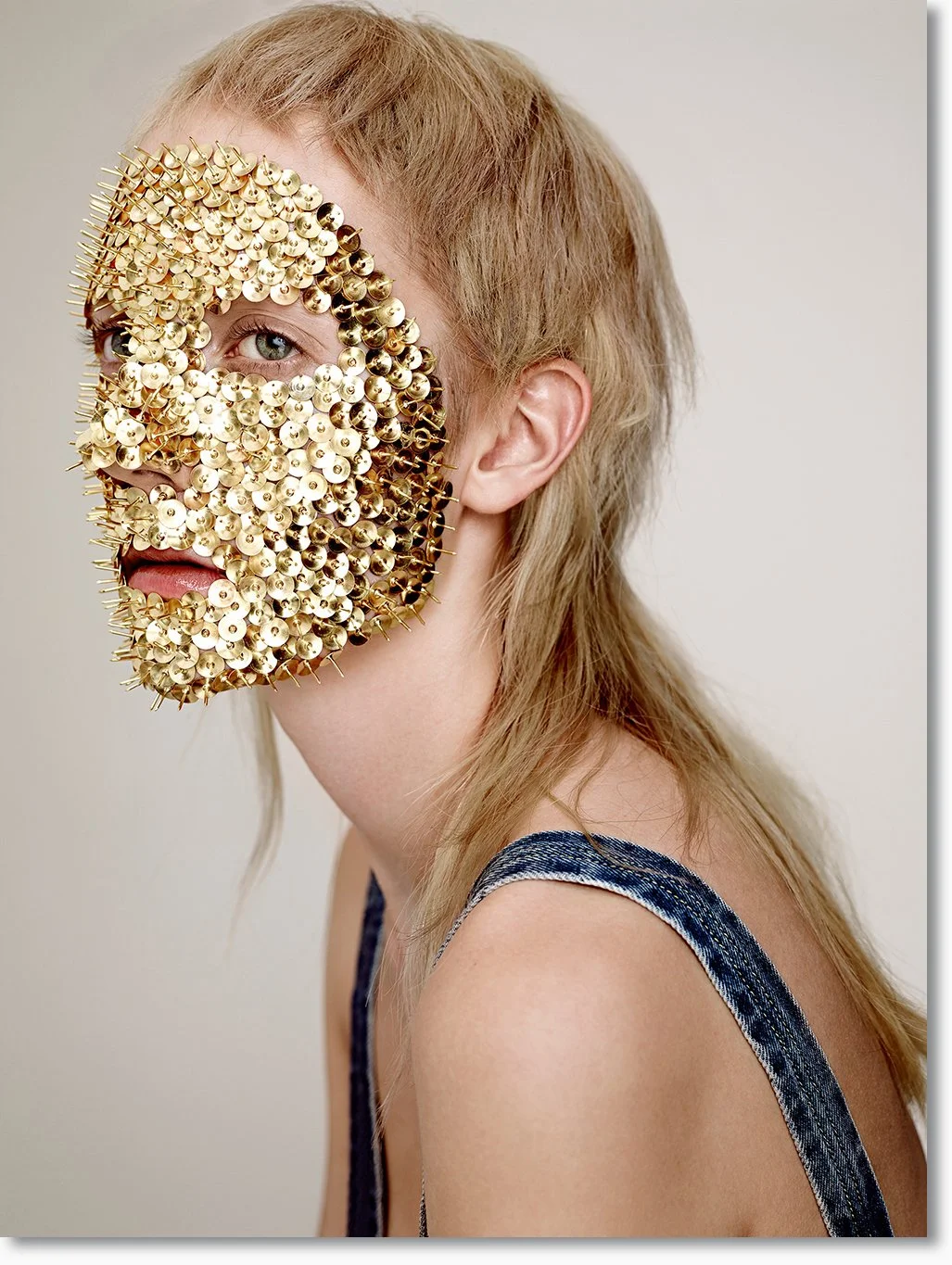 Eine Frau mit blonden Haaren trägt eine künstlerische Maske aus goldfarbenen Reißzwecken, die ihr halbes Gesicht bedeckt.