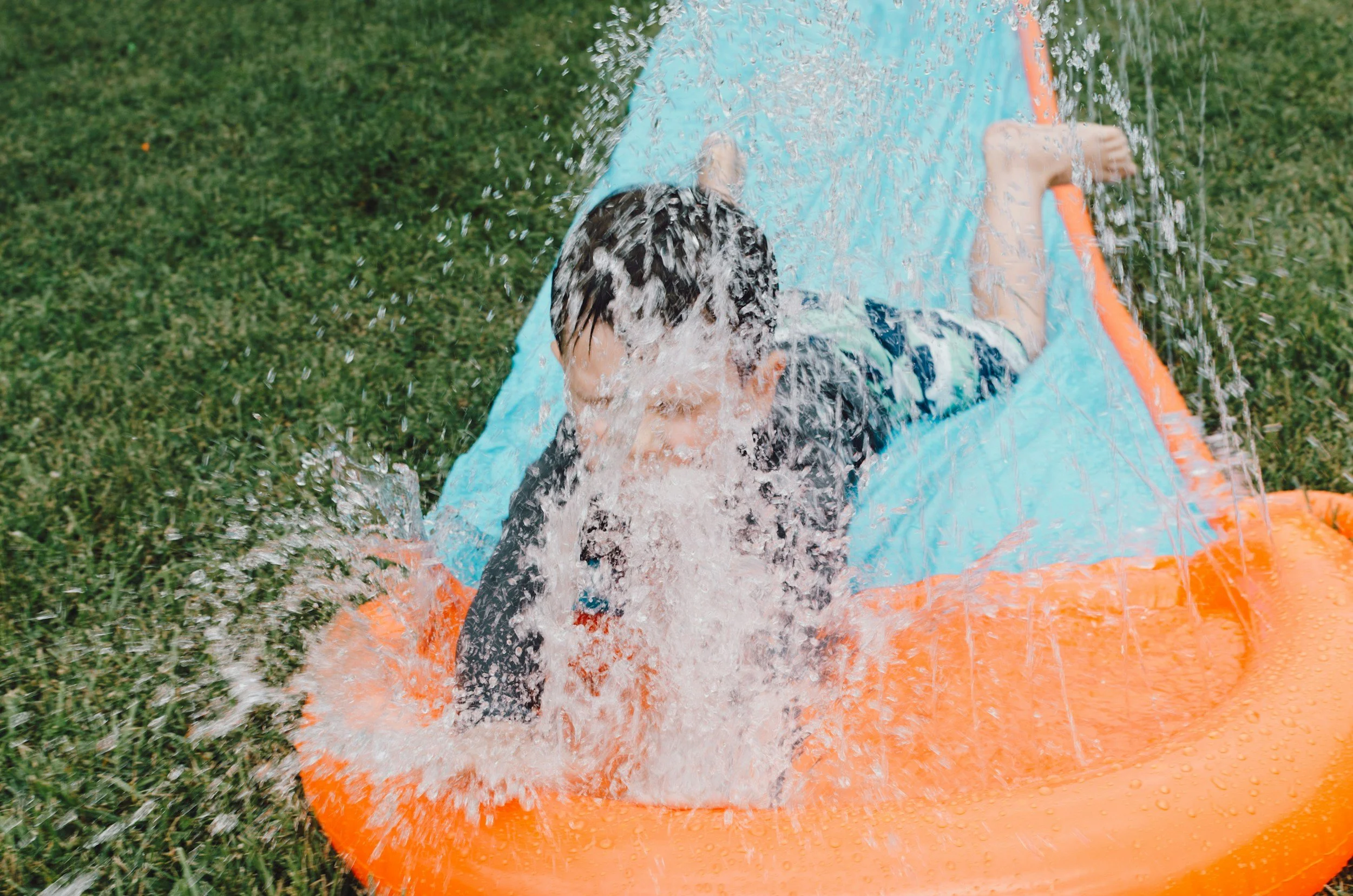 Child playing on an orange and blue inflatable water slide outdoors, spraying water over his face on a grassy area.