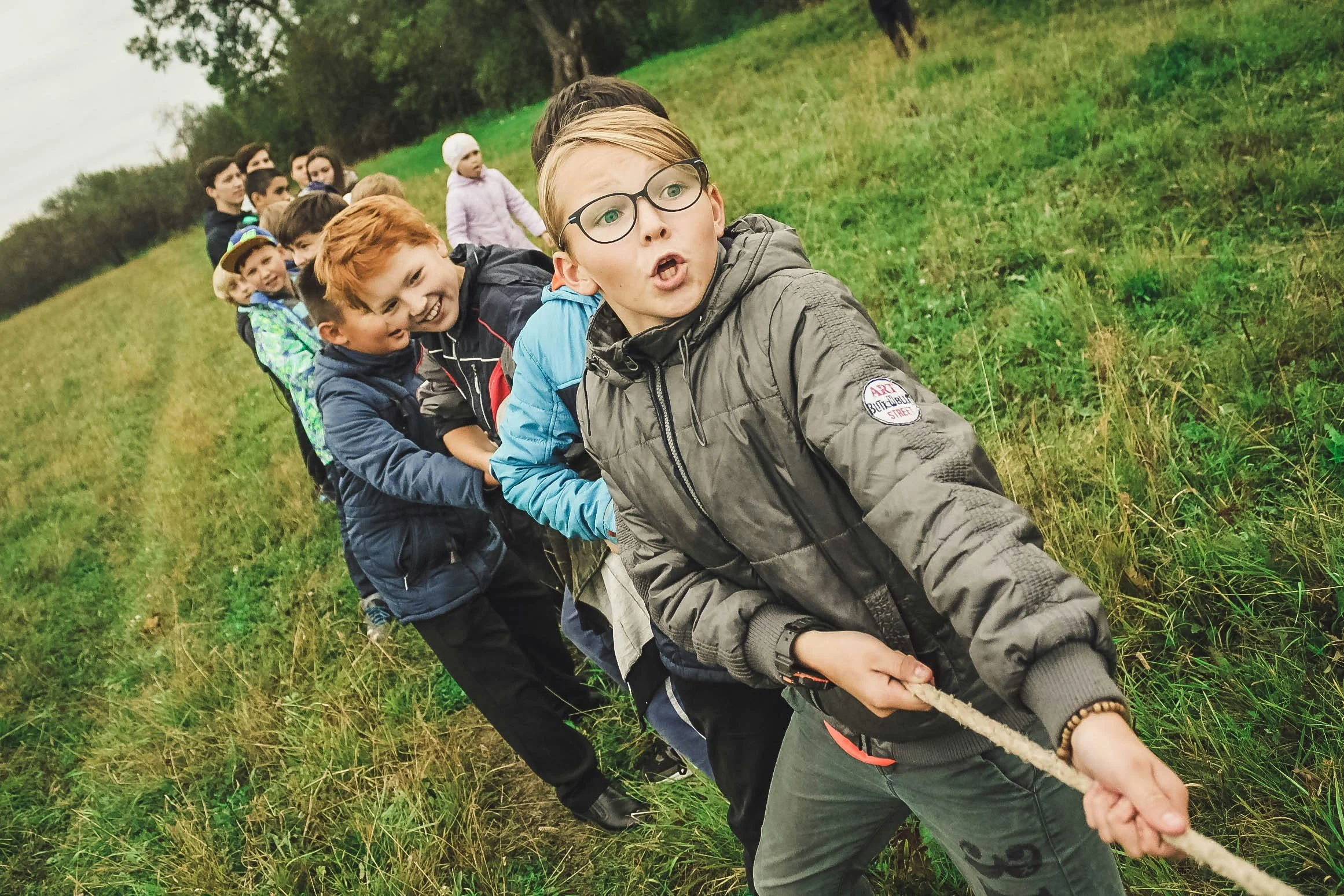A group of children are participating in a tug-of-war game outdoors on a grassy field. The boy at the front wearing glasses and a grey jacket is pulling the rope, making an exaggerated facial expression. Other children are lined up behind him, holding the rope tightly and smiling, with some looking excited and others focused. The setting appears to be a cloudy day in a rural or park area.