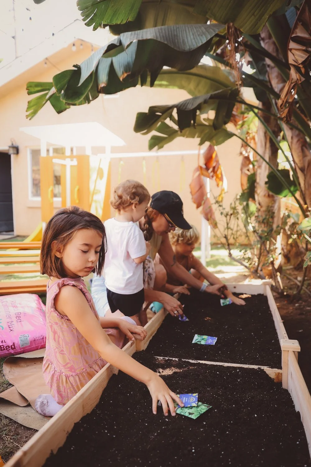 Children and adults planting seeds in a garden bed in a backyard, with tropical plants and a house in the background.