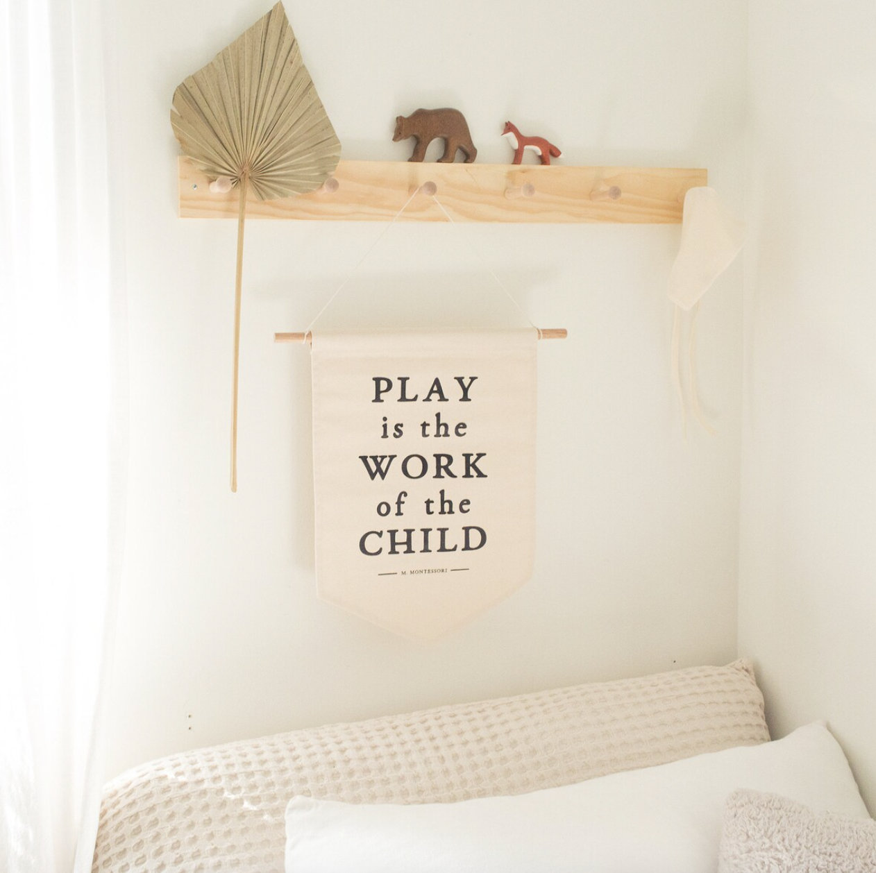 Decorative wall with a hanging banner that says, 'Play is the work of the child' by M. Montessori. Above the banner is a wooden shelf with small animal figurines and a fan-shaped decorative item.
