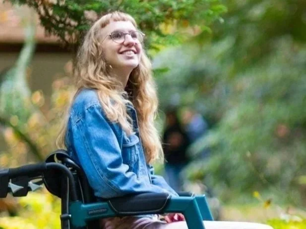 A blonde woman in a power wheelchair smiles up at the trees in a park