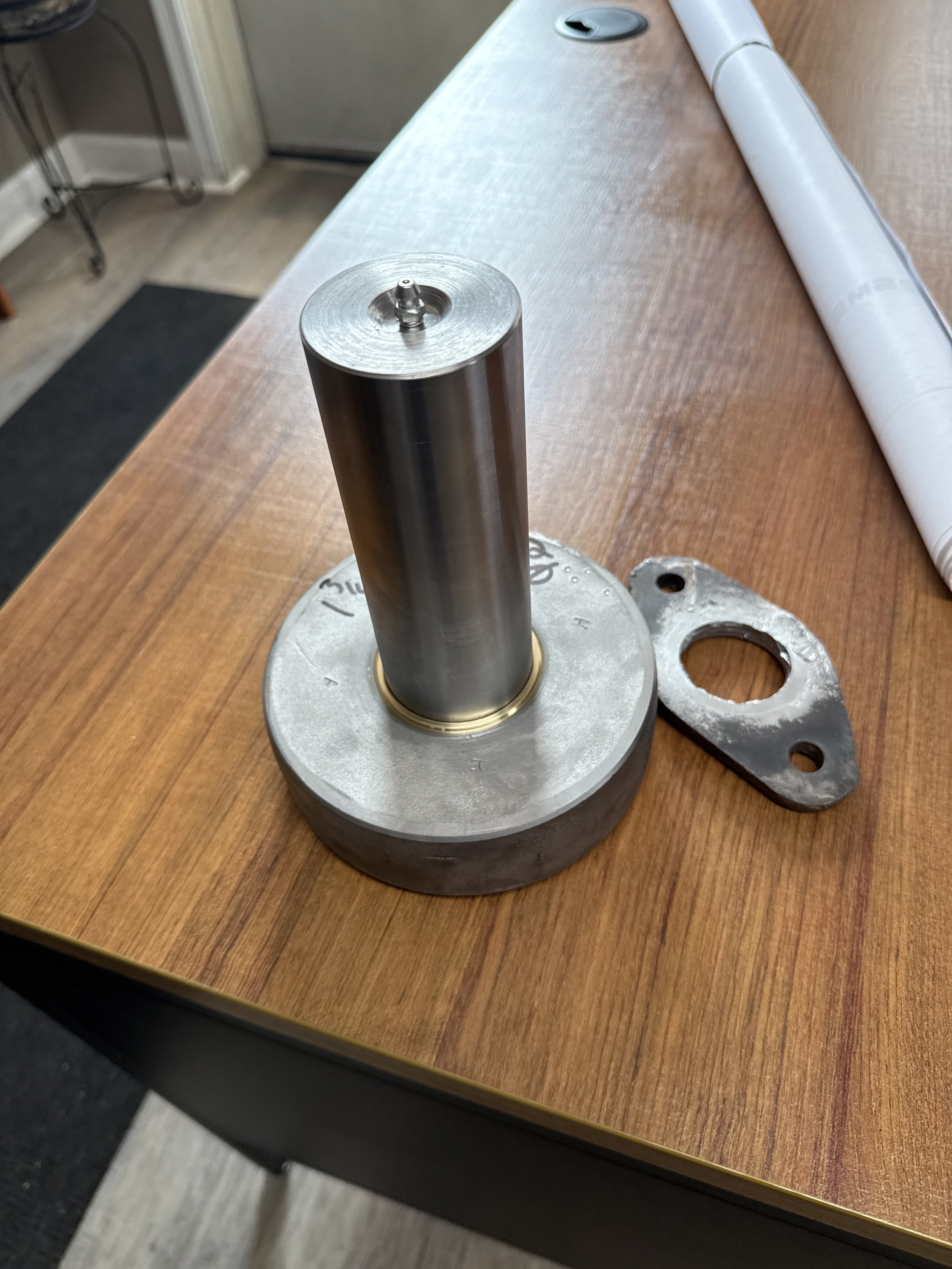 Metal machine part with a large cylindrical shaft, set on a wooden table. A metal gasket or plate with holes for bolts is next to it, and a rolled-up blueprint is partially visible.