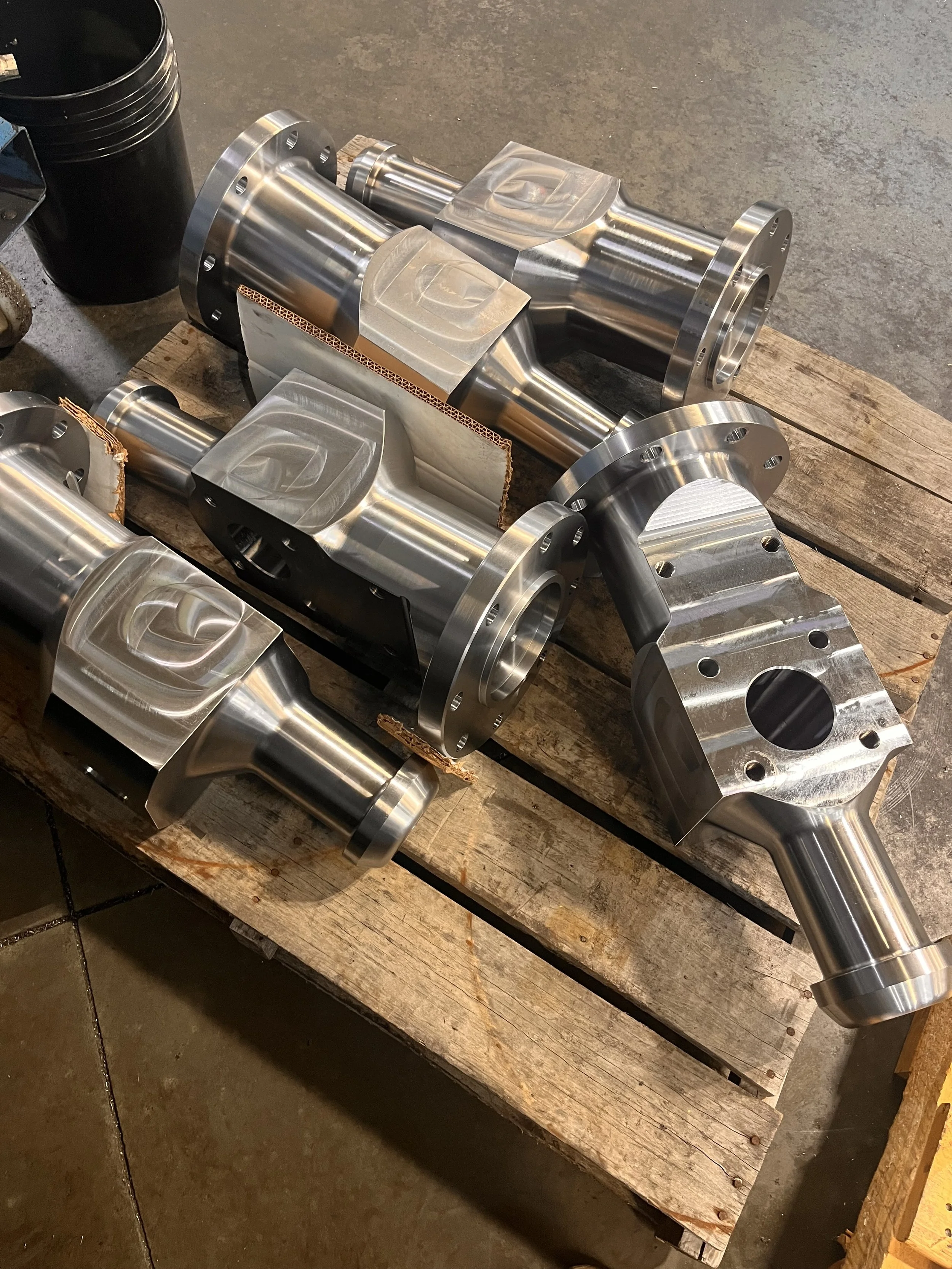 Three large, shiny metal machined automotive parts on a wooden pallet in a workshop.