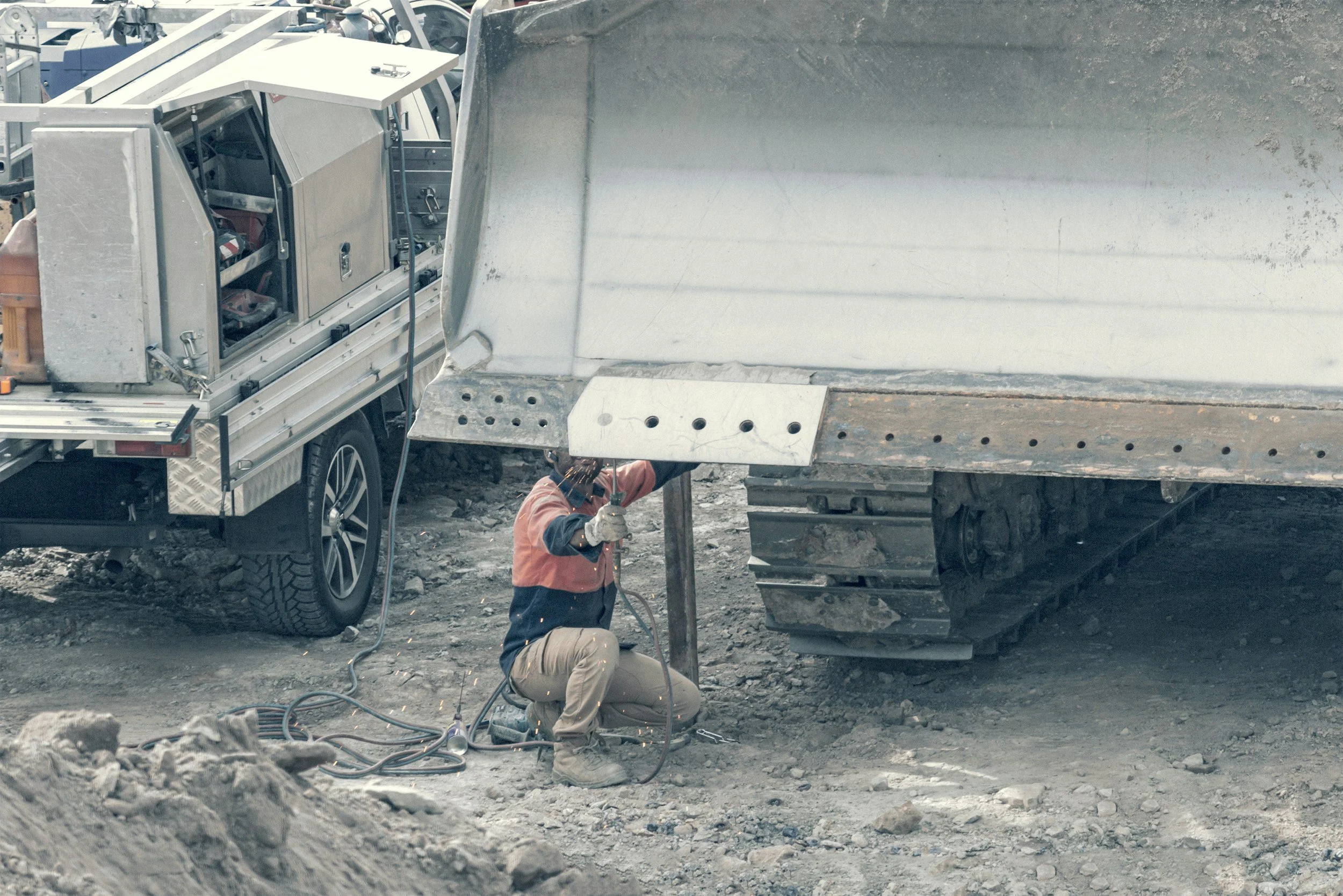 A worker in safety gear welding a large metal structure at a construction site, with a metal truck and a service vehicle nearby.