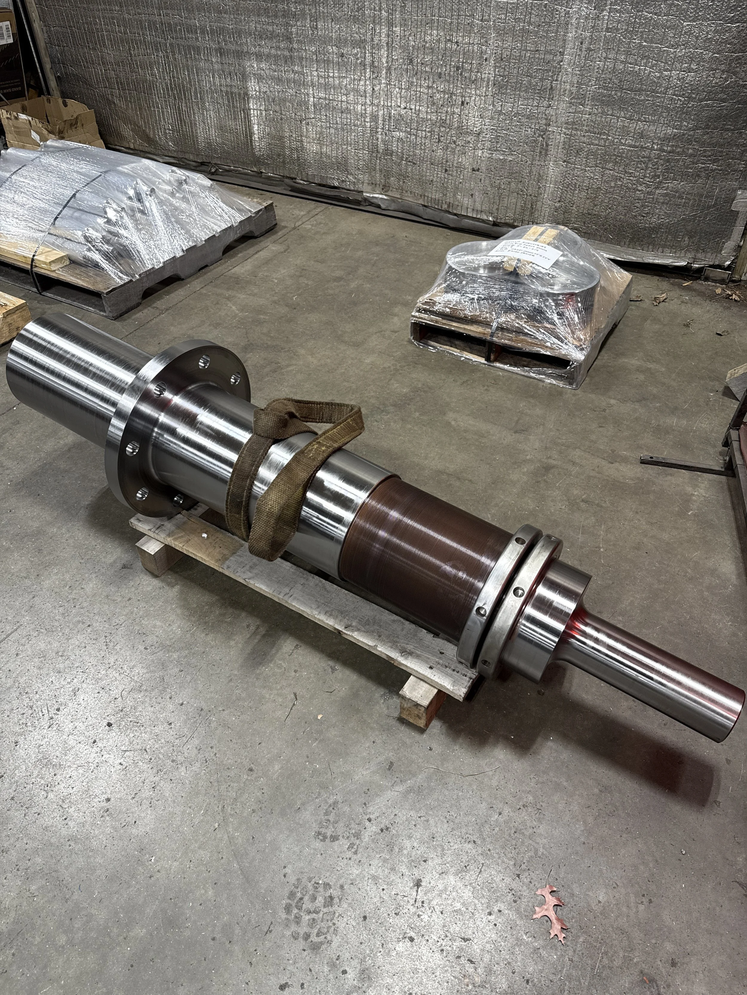 Large metal industrial shaft resting on wooden pallets in a warehouse, with wrapped packages and insulation material in the background.