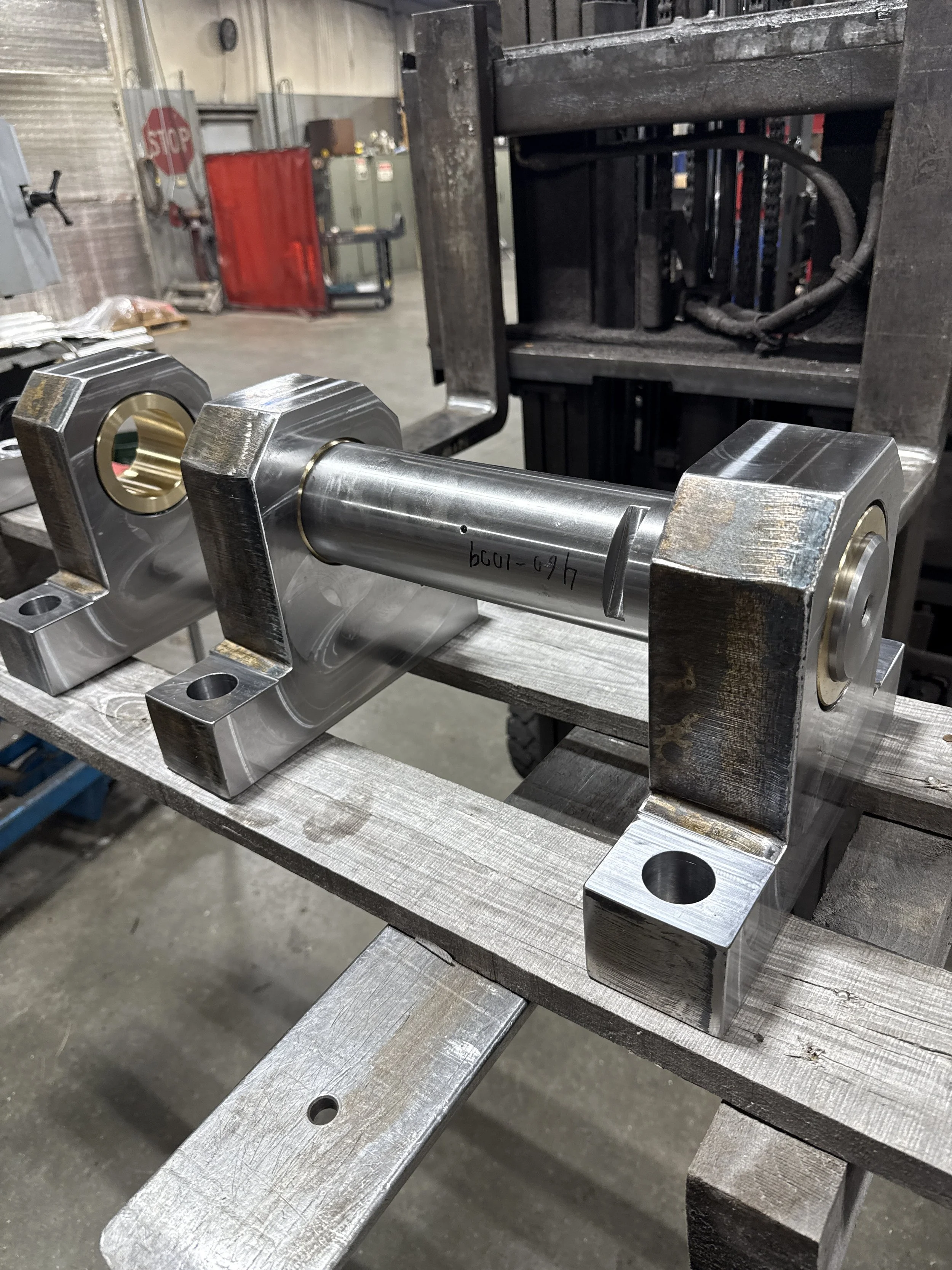 Metal component with a cylindrical shaft and two block-shaped ends mounted on a workbench in an industrial workshop.