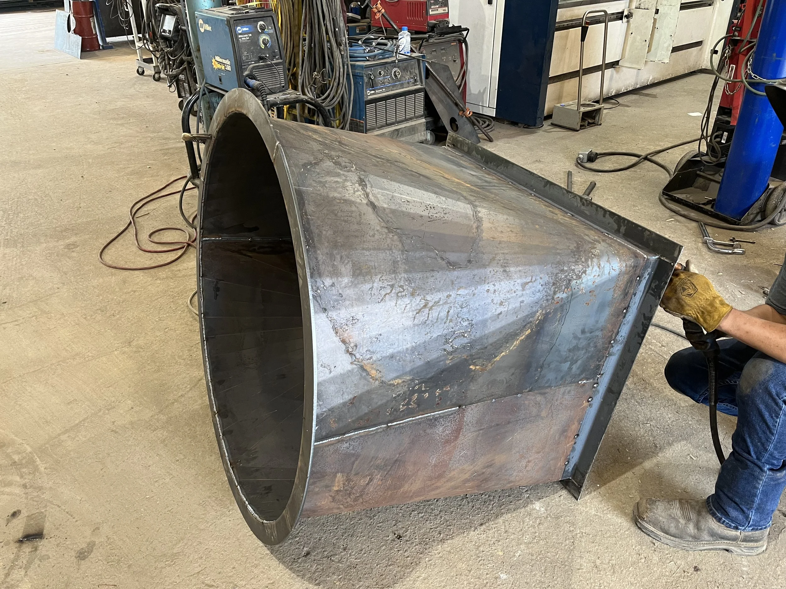 A person welding a large, cylindrical metal structure in an industrial workshop. The structure is made of steel and appears to be part of a larger project, with welding equipment and tools visible in the background.