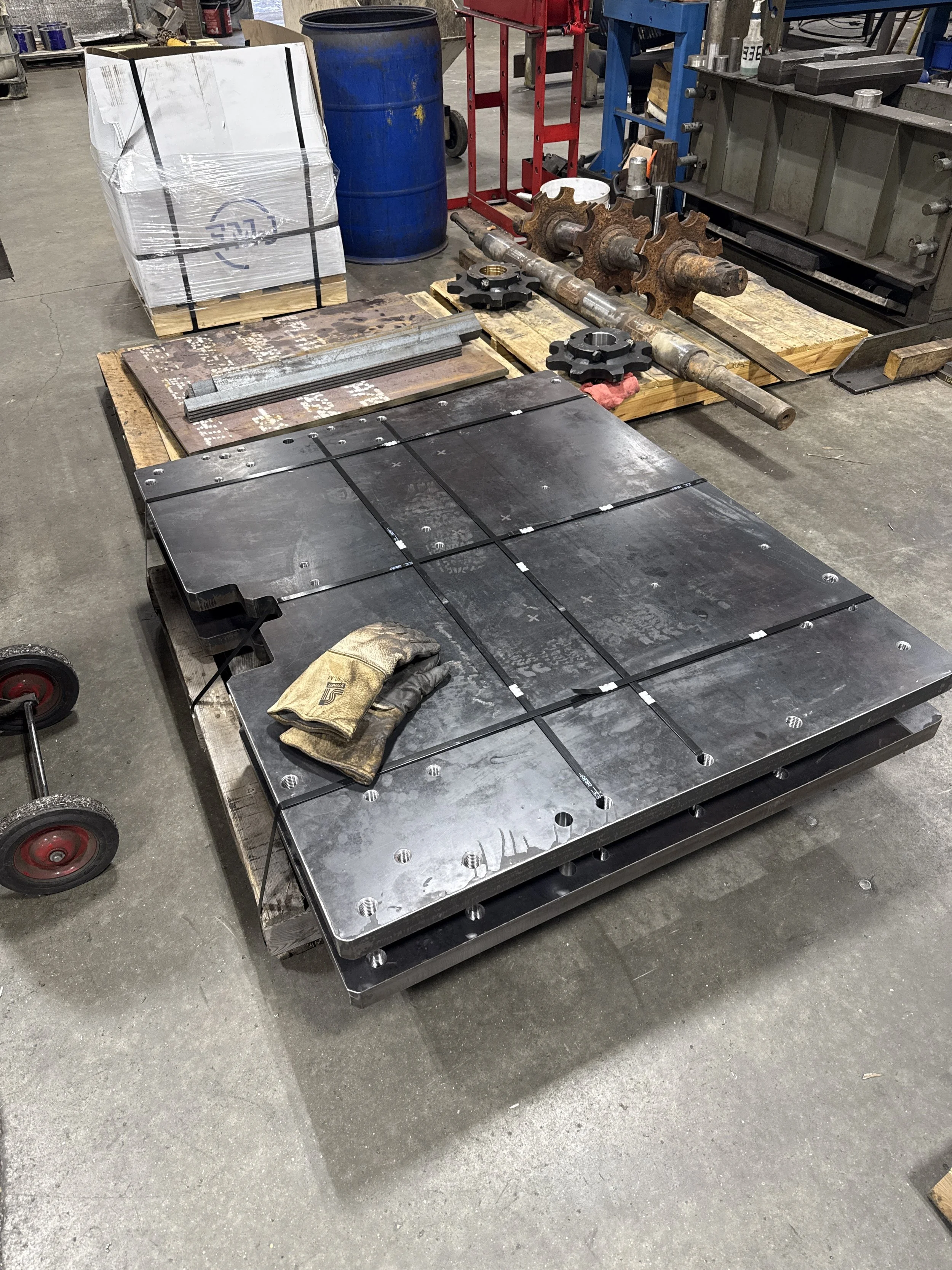 Large metal plate with multiple smaller plates attached on top, placed on a wheeled cart in an industrial workshop. Gloves resting on the surface, with tools and machinery around.