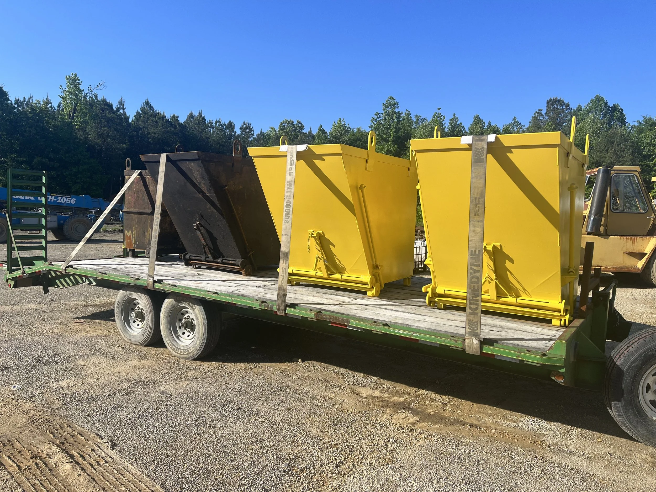 A flatbed trailer carrying two yellow and one black waste containers on a gravel lot, with trees and a blue sky in the background.