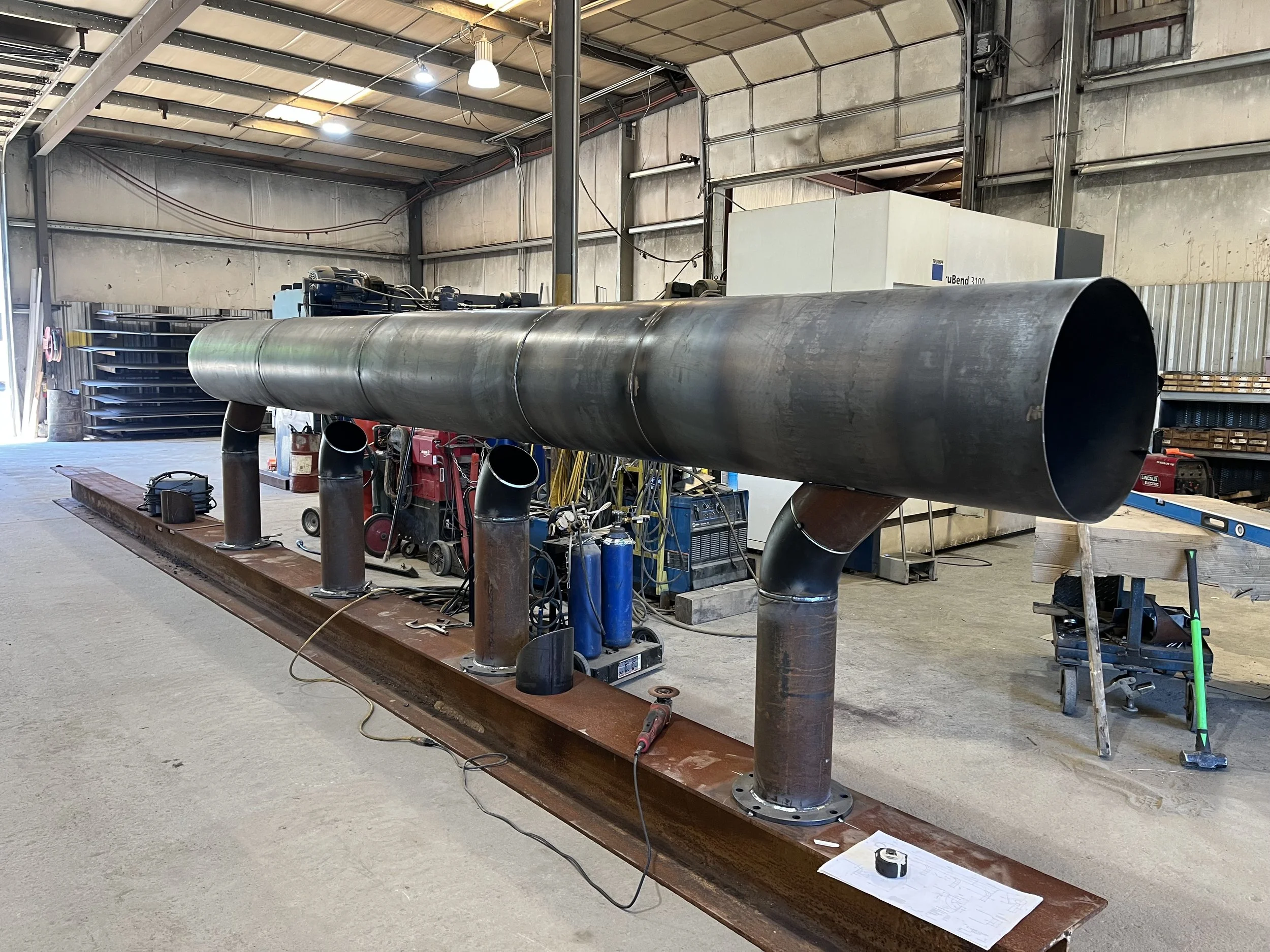 Large industrial pipe with attached smaller pipes and fittings, supported on a metal frame inside a warehouse or workshop, with tools and equipment around.