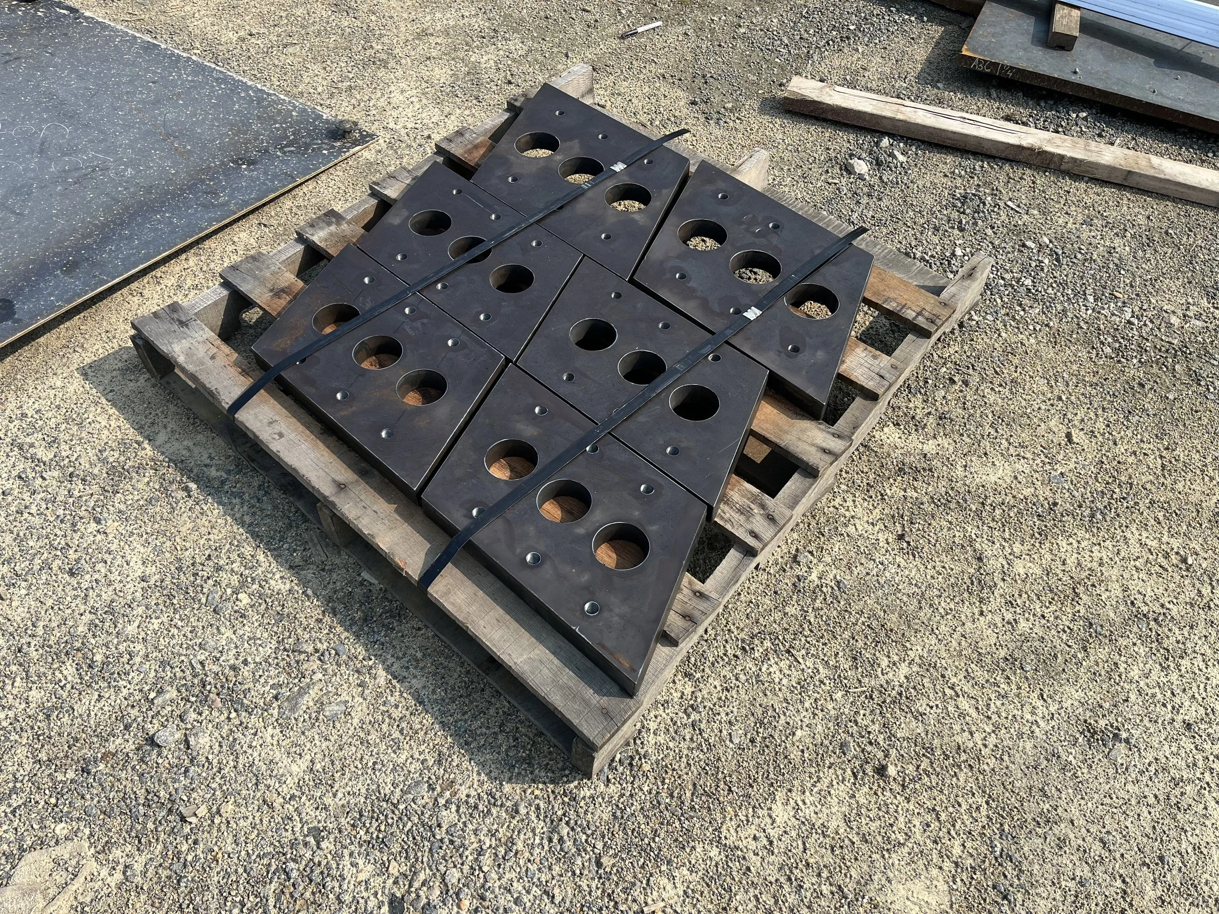Eight black metal plates with holes stacked on a wooden pallet outdoors.