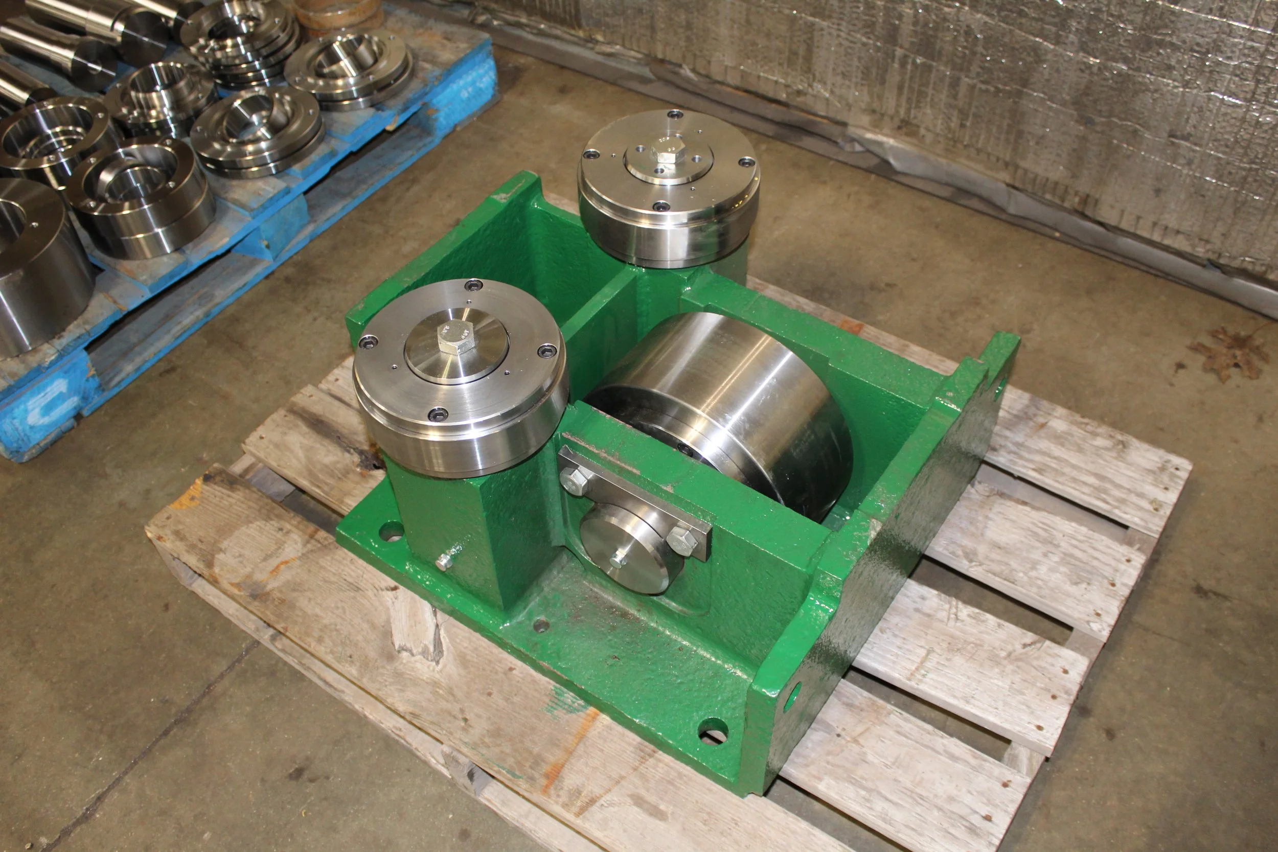 Industrial machinery component with green painted metal base and several shiny metal rollers or wheels, on a wooden pallet in a workshop.