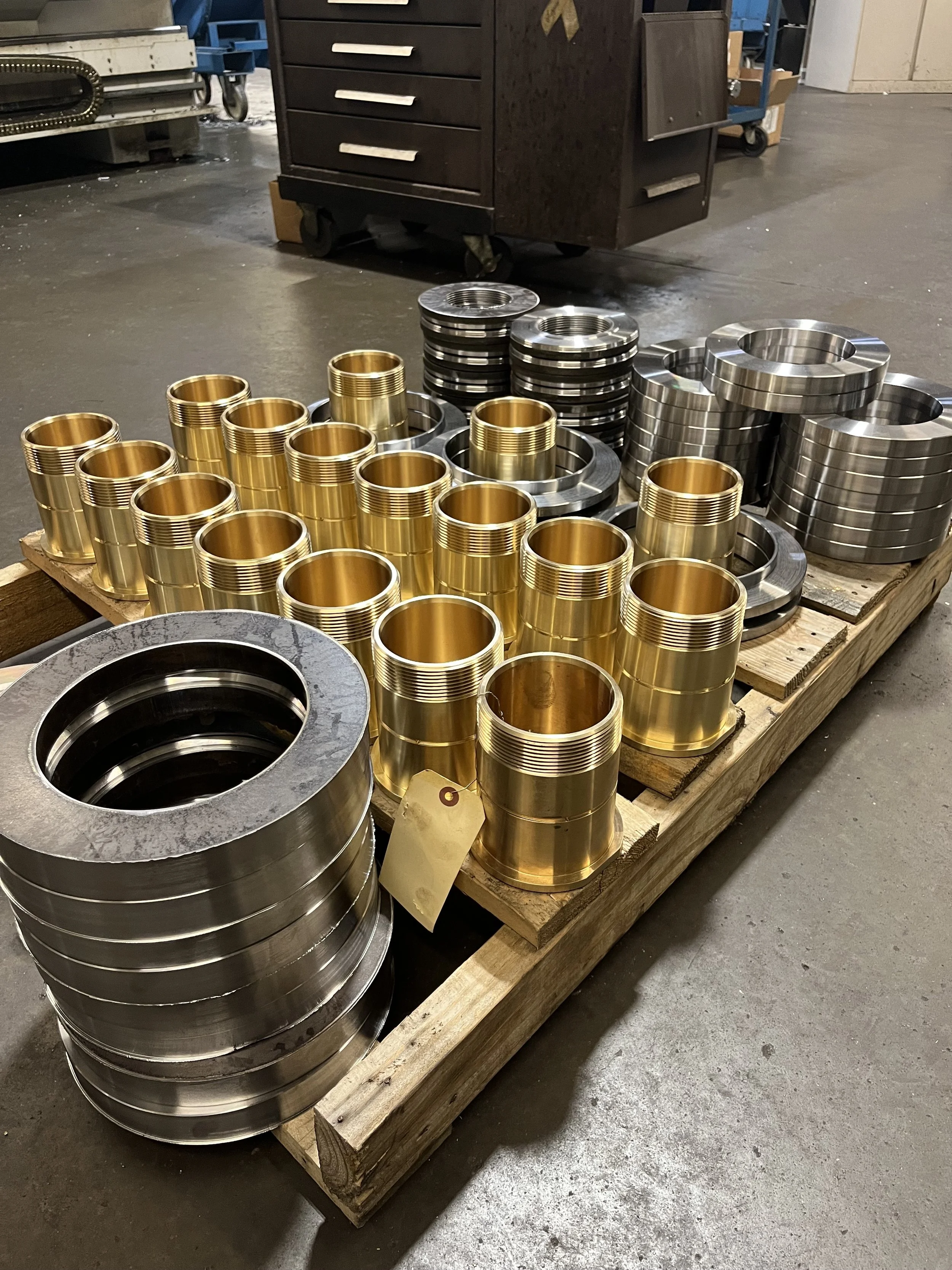 Metal machine parts in gold and silver, arranged on a wooden pallet in an industrial setting.