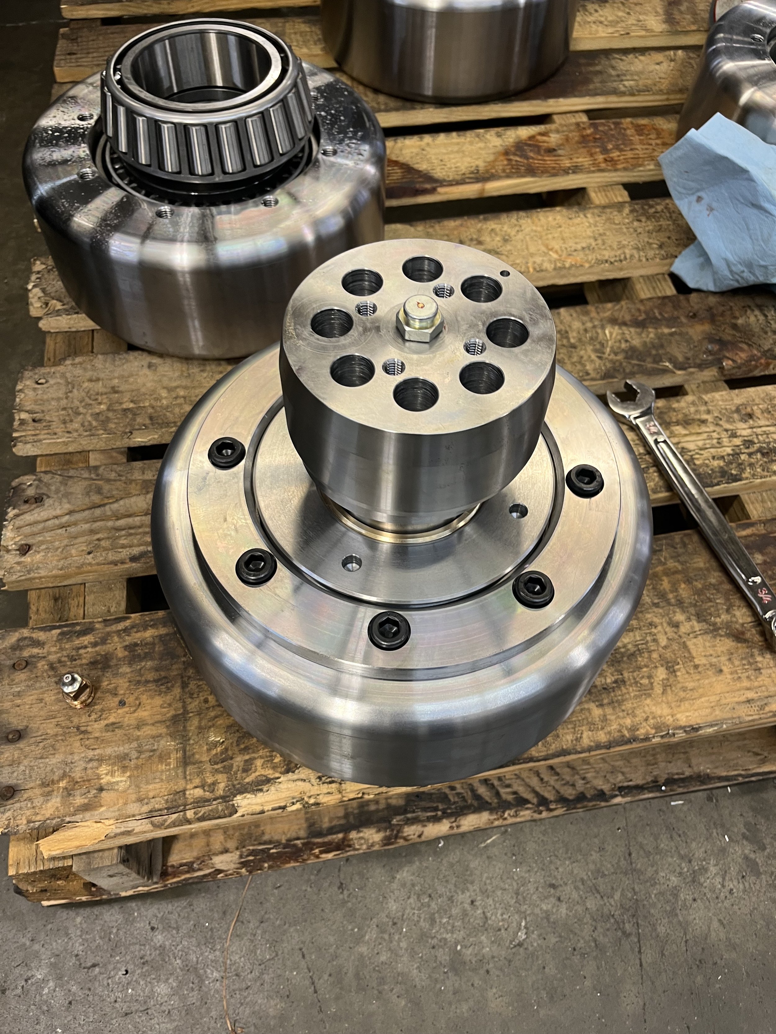 Close-up of a large, shiny metal mechanical component with multiple circular holes, bolts, and black screws, placed on a weathered wooden workbench with tools and a paper towel nearby.