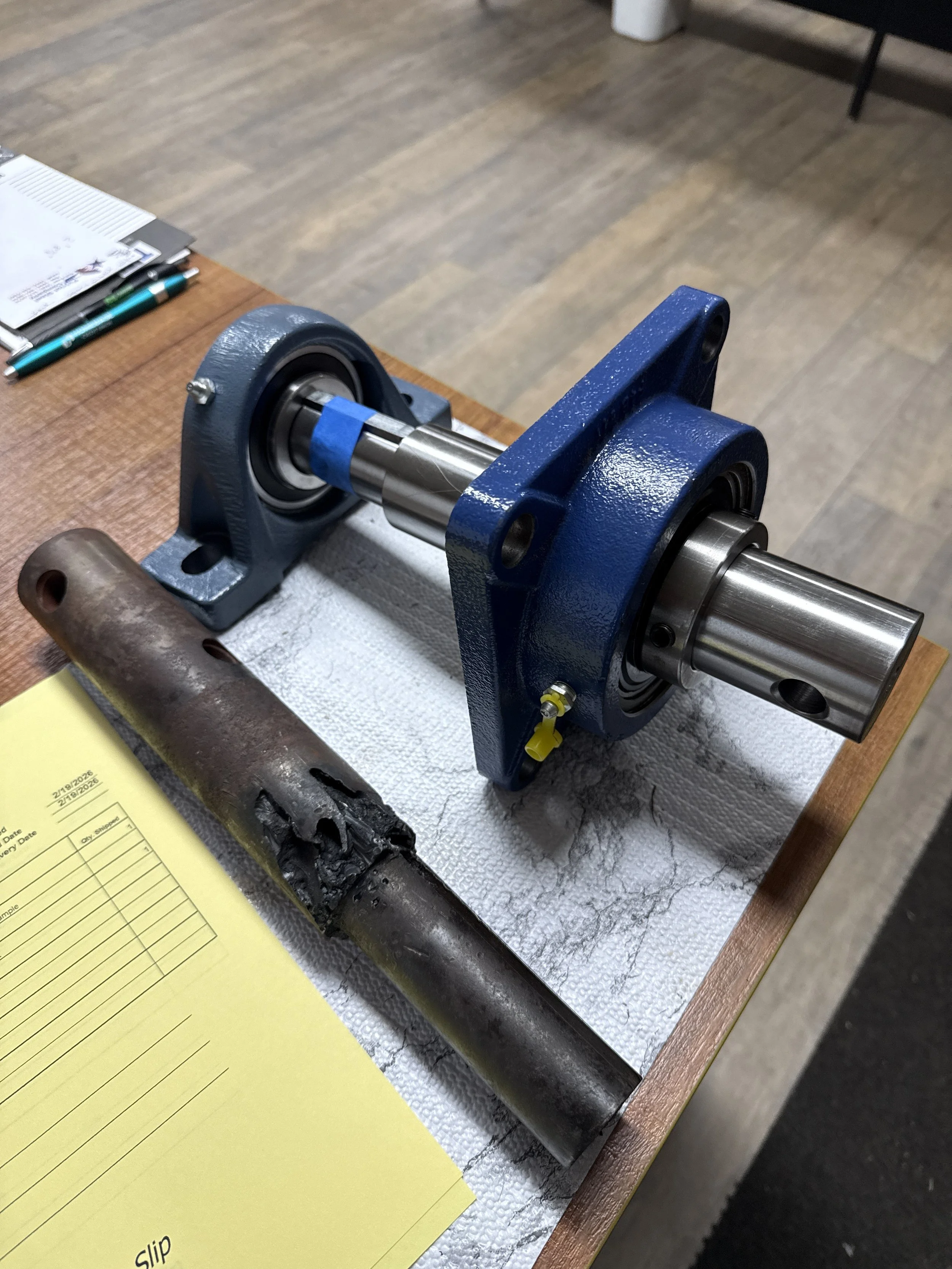 A blue mounted bearing housing with a metal shaft inserted, next to a rusted pipe on a workbench.