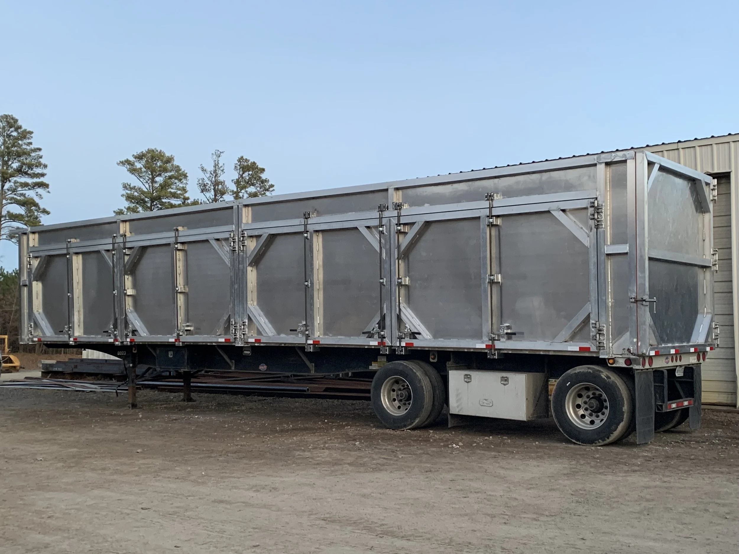 A large metallic livestock trailer parked on a dirt ground near a building with trees in the background.