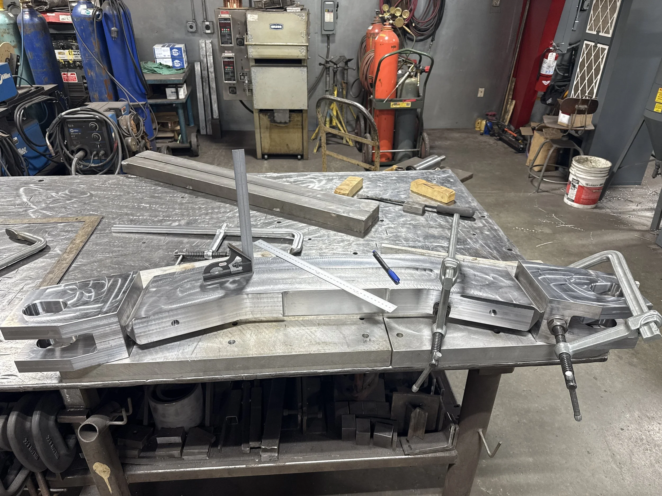 Metalwork workshop with a large metal piece secured on a workbench, surrounded by clamps, straightedges, and various tools.