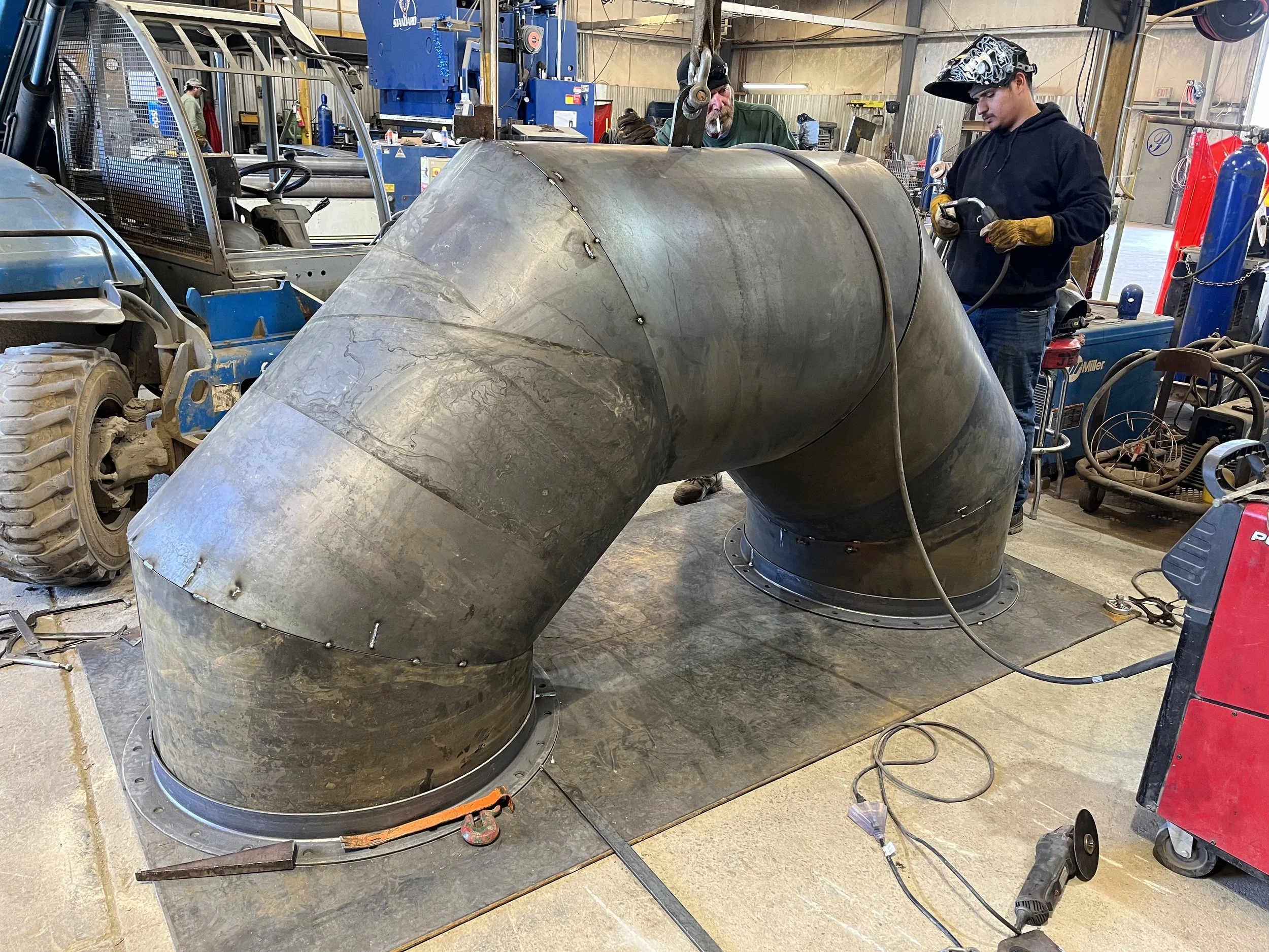 Two workers welding or working on a large metal pipe or duct in a workshop. The pipe is bent and secured with bolts, with welding equipment nearby.