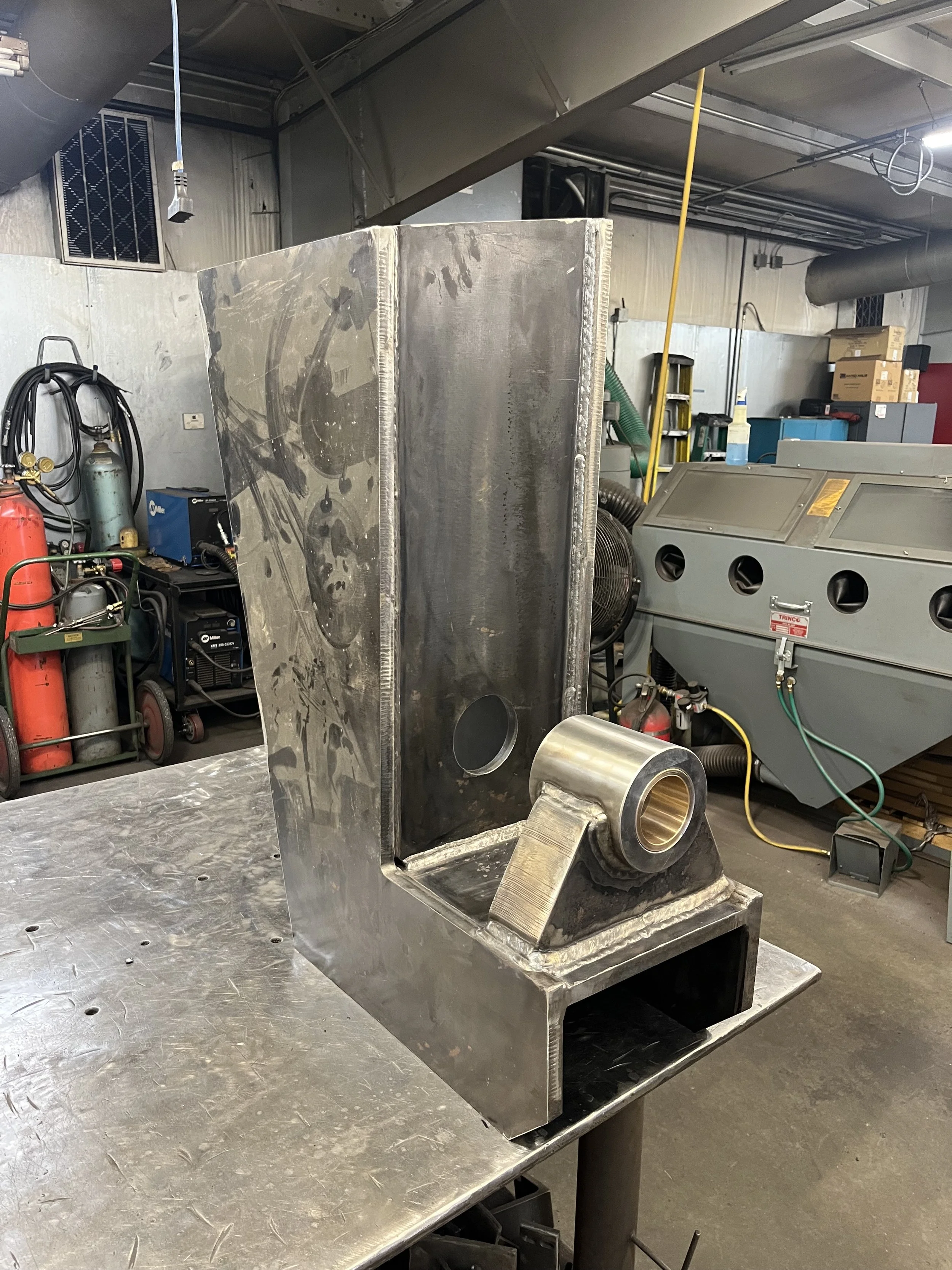 Metal fabrication in progress with a large, rectangular, welded stainless steel component on a workbench, featuring a circular opening and a welded pipe joint. Workshop tools and equipment are in the background.