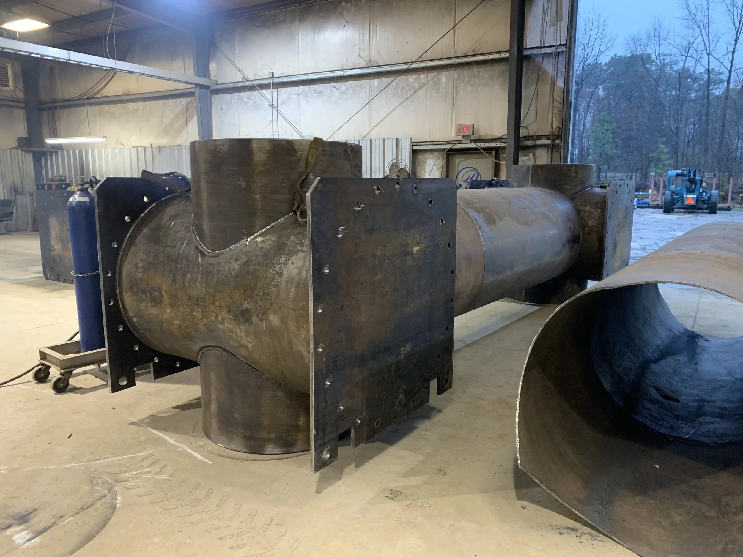 Large industrial metal pipes in a warehouse, with a large inside pipe and some fittings and equipment nearby.