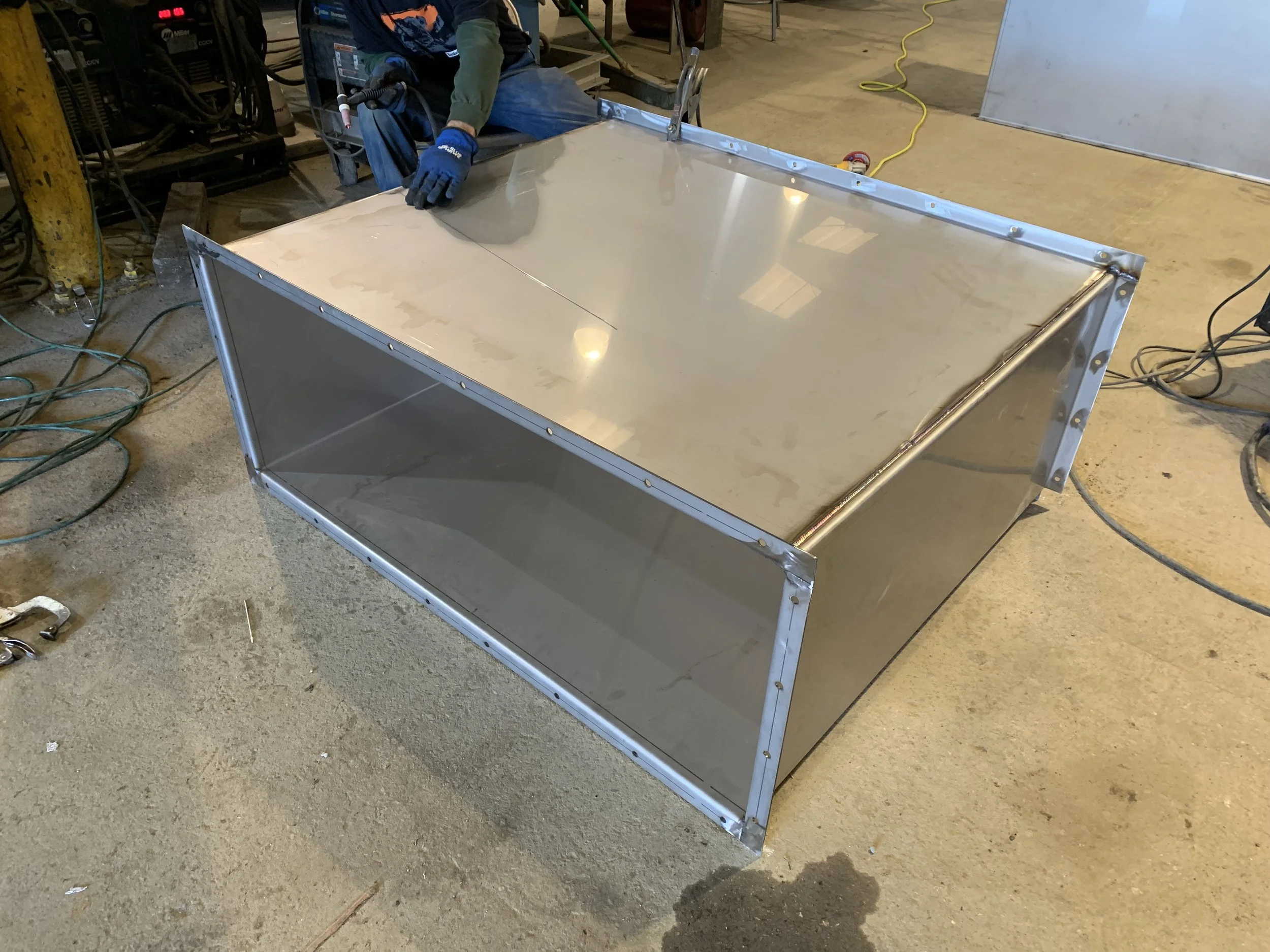 A worker in a workshop installing or assembling a large metallic structure or enclosure, with tools and wires around, on a concrete floor.