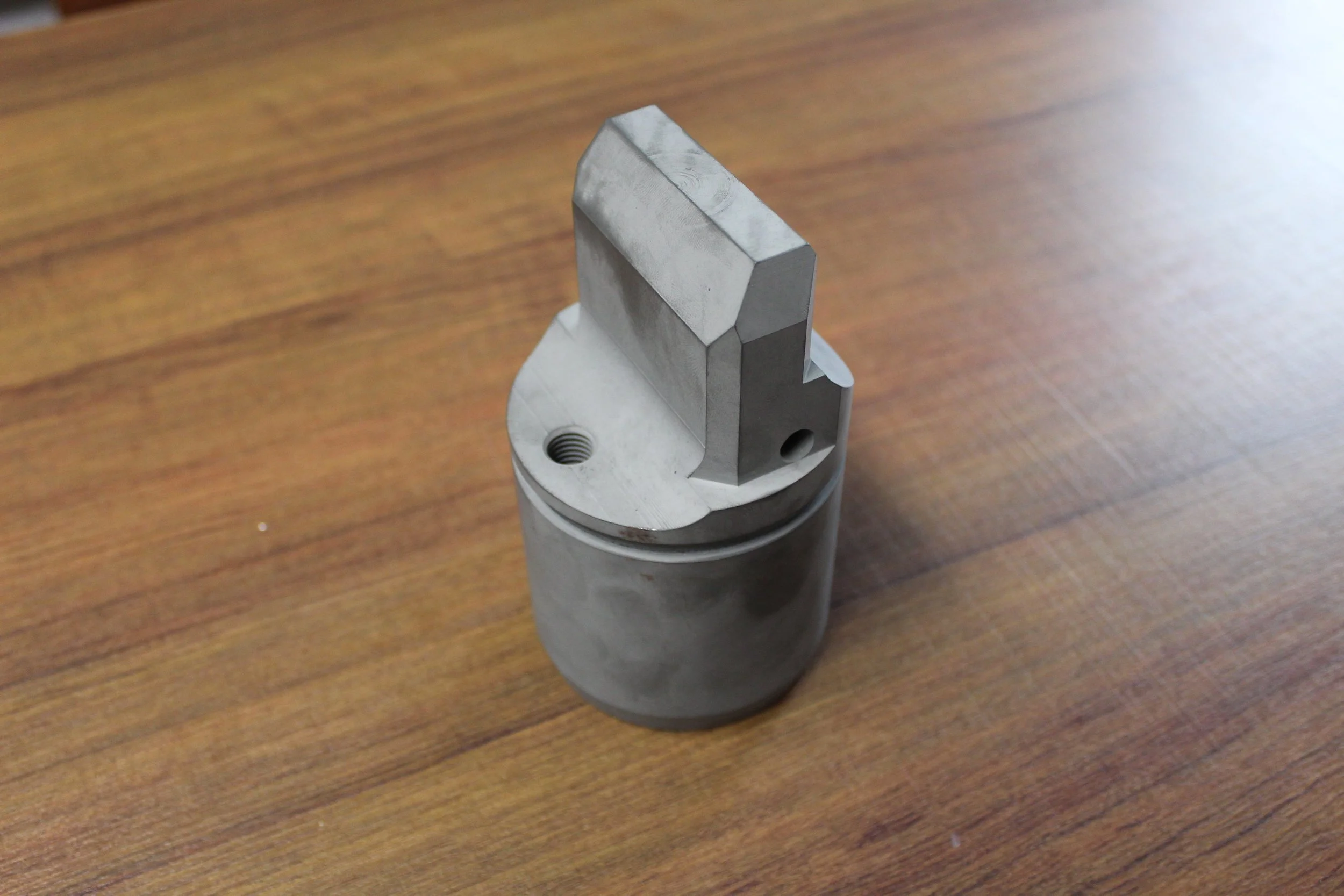 A metallic industrial part with a cylindrical base and a rectangular protrusion on top, placed on a wooden surface.