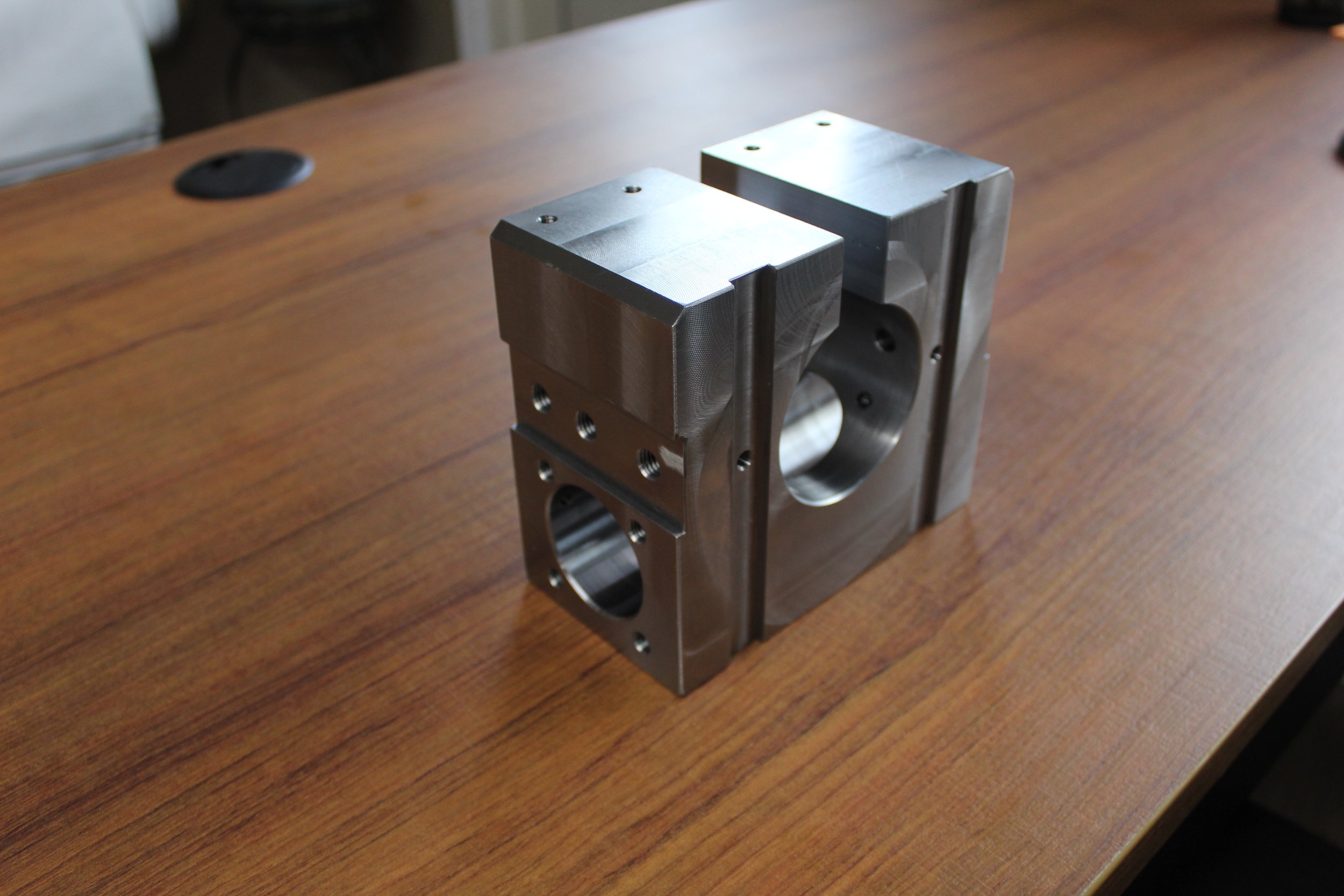 Metallic mechanical part on a wooden table with cylindrical and rectangular shapes, multiple holes, and grooves.