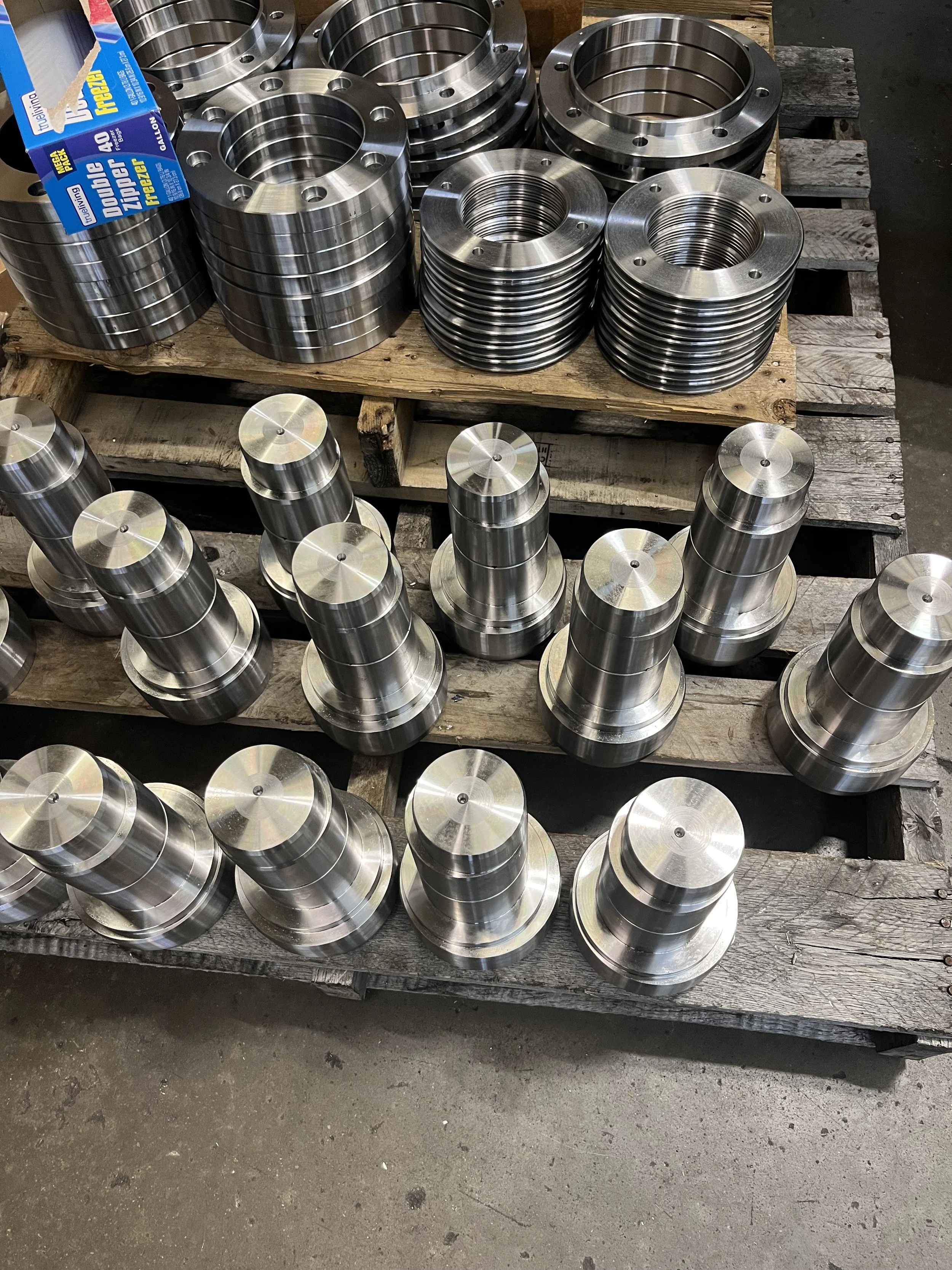 Metal machined parts arranged on wooden pallets, including cylindrical components with flares and holes, likely for industrial purposes.