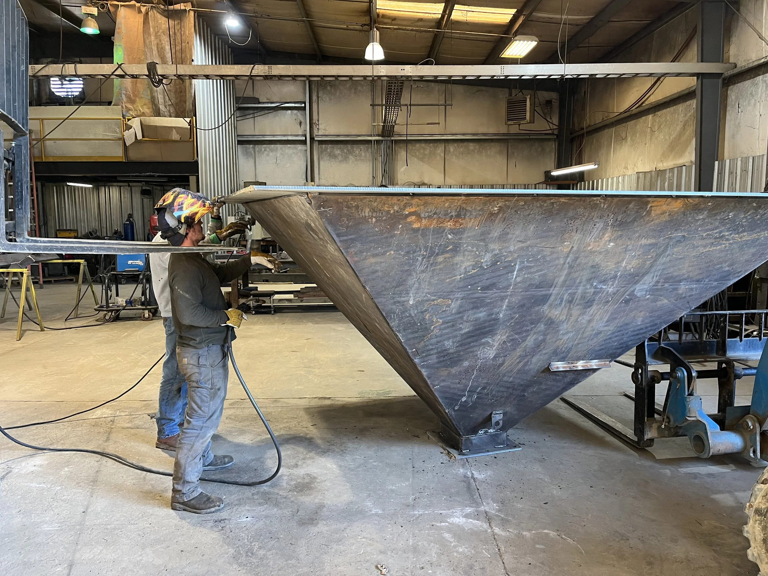 Two workers welding large metal structure inside a workshop. One worker is wearing a colorful helmet, gloves, and protective clothing, holding a welding tool while working on the metal piece. The workshop has industrial equipment and tools in the bac