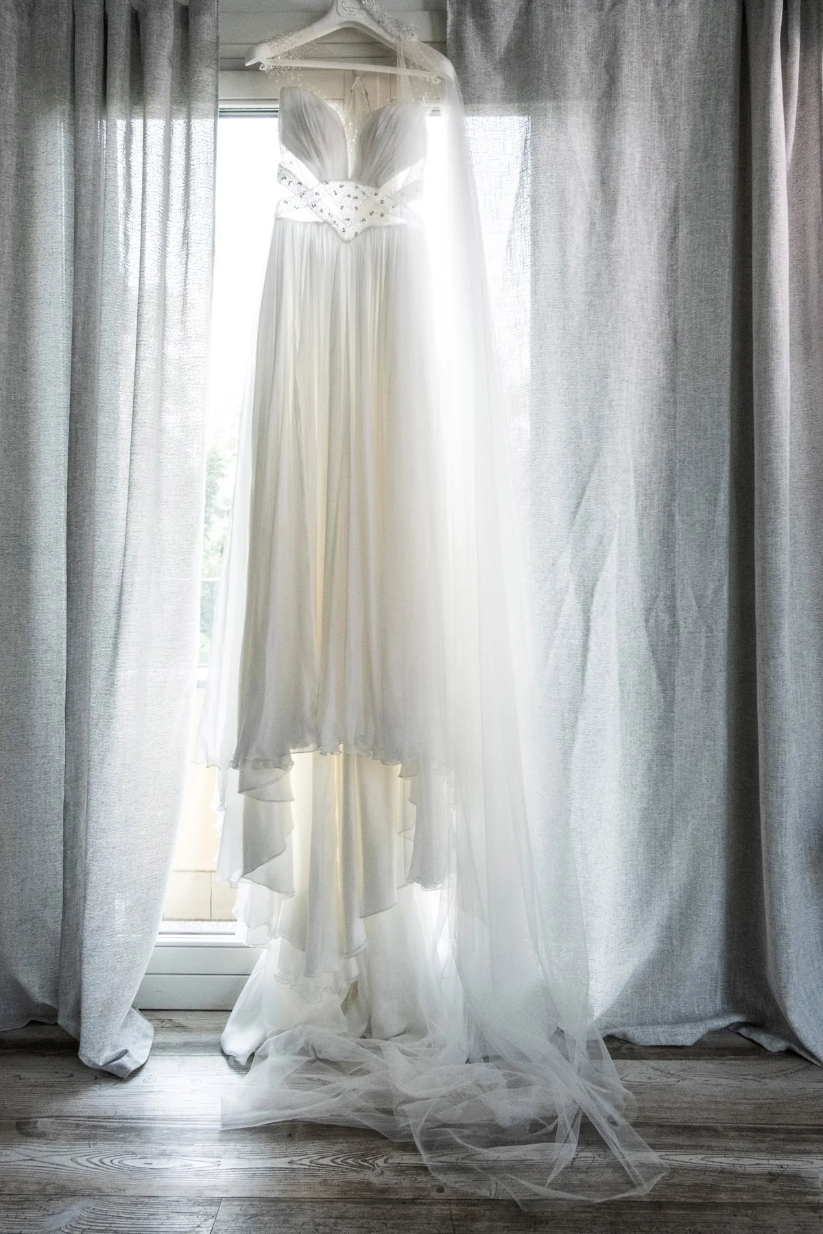 Abito da sposa bianco appeso tra le tende di lino grigio chiaro, con luce che filtra dietro. Wedding planner Varese.
