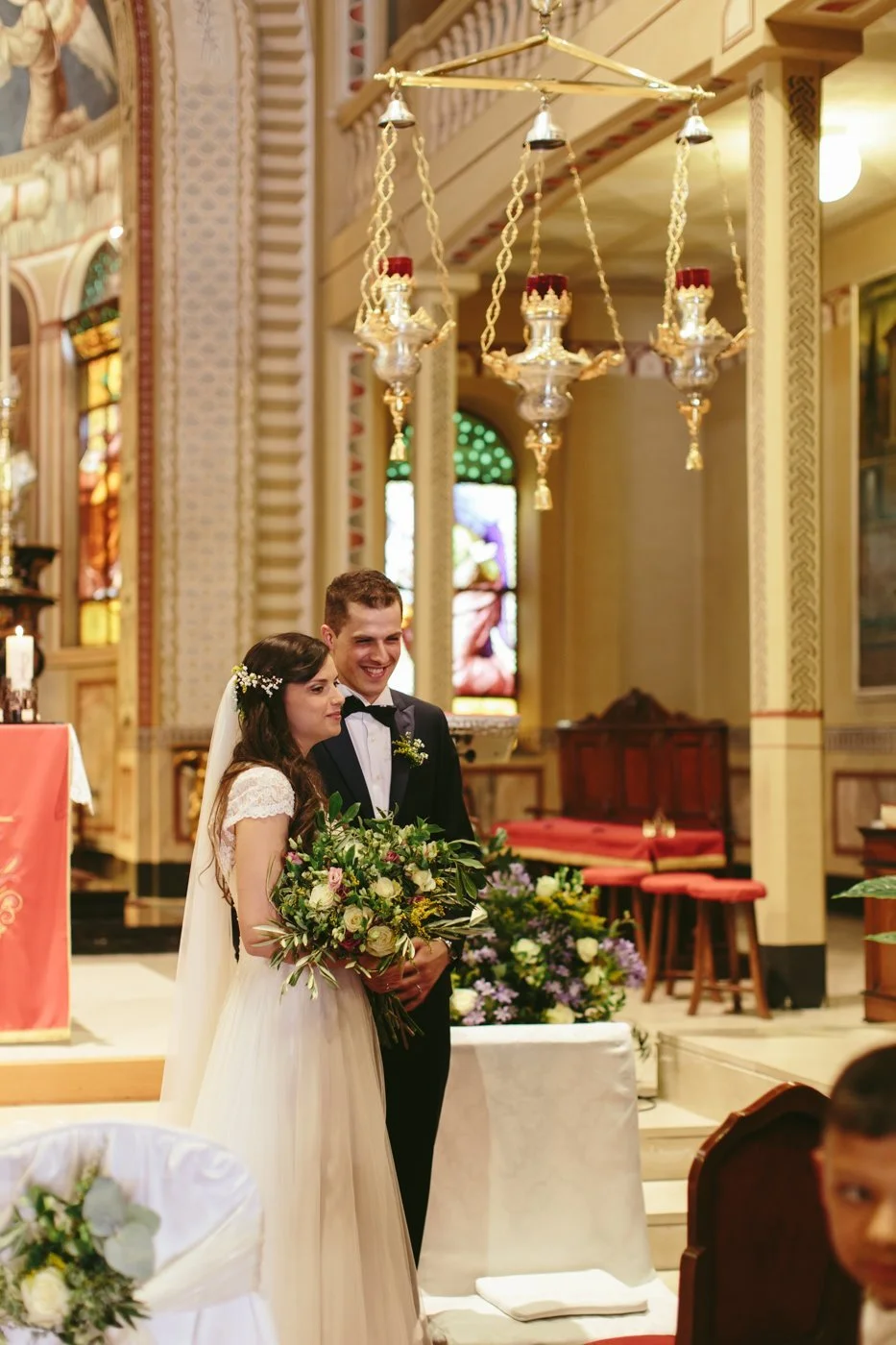Sposi in chiesa durante il matrimonio, con il sposo sorridente e la sposa che tiene un mazzo di fiori, ambientazione sacra con vetrate colorate.
