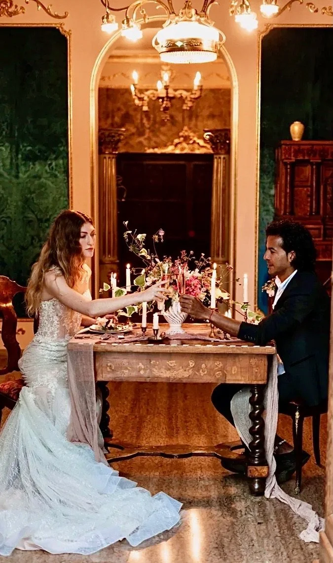 Una coppia innamorata si scambia un brindisi durante una cena elegante in una sala decorata con candelabri e un grande specchio.