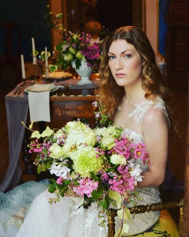 Una donna con capelli mossi porta un abito bianco da sposa e tiene un grande bouquet di fiori colorati in un ambiente che sembra una sala da pranzo con decorazioni floreali.