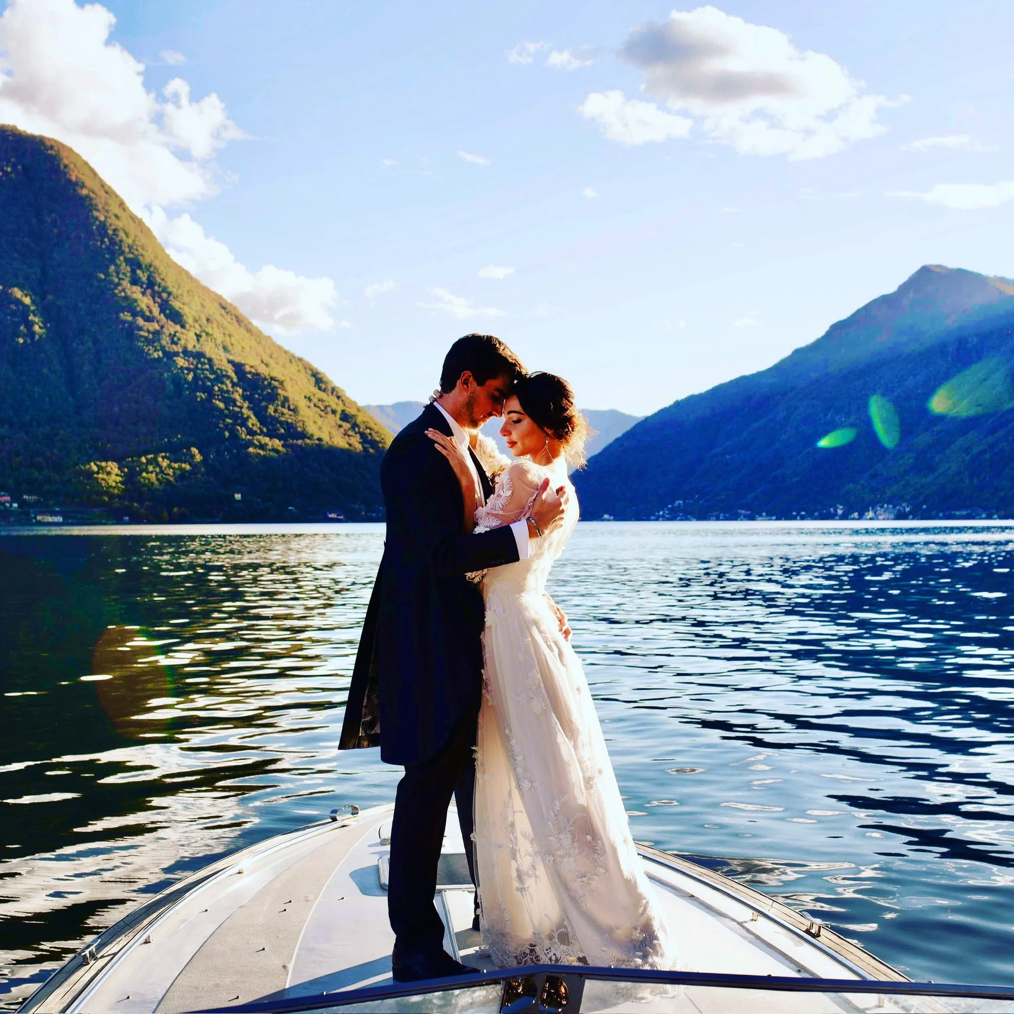Una coppia di sposi si abbraccia e si guarda negli occhi su una barca in piedi su un lago con montagne sullo sfondo, al tramonto.