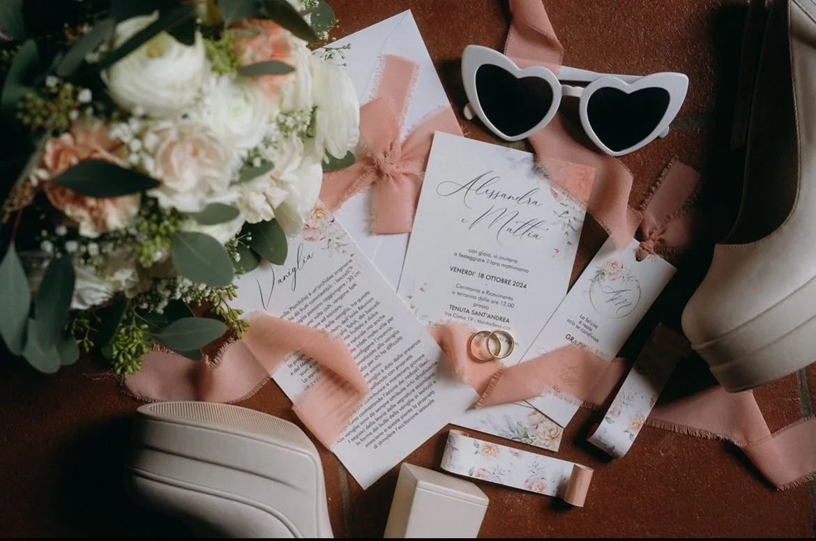 Invito di nozze con bouquet di fiori, occhiali da sole a forma di cuore, due fedi nuziali, e alcuni oggetti da sposa come scarpe e abito, disposti su un tavolo di legno.