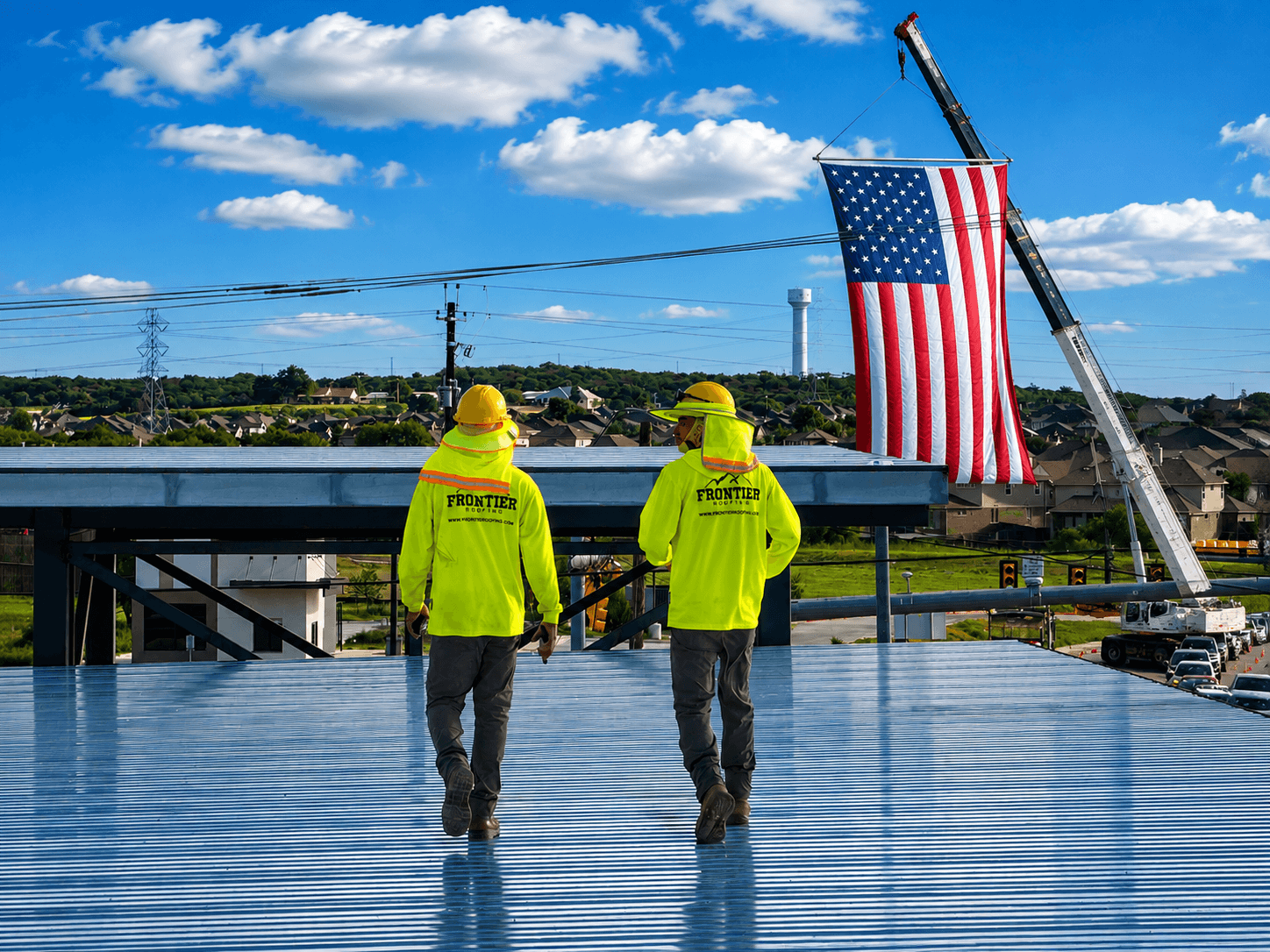 Two workers in yellow safety jackets and helmets walking on a rooftop with a large American flag hanging from a crane behind them.