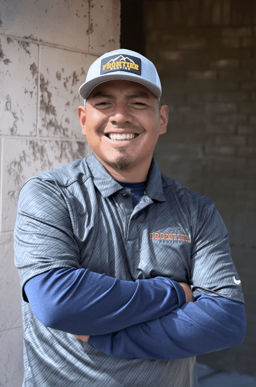A man smiling with crossed arms, wearing a baseball cap and a gray and blue Frontier Roofing shirt, standing in front of a textured wall.
