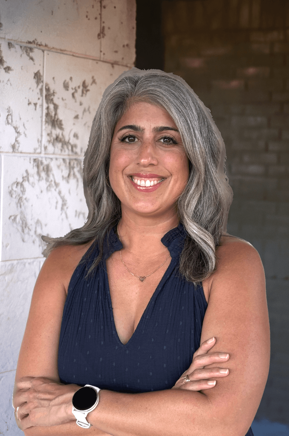 A smiling woman with gray hair, wearing a navy blue sleeveless top, a necklace with a heart pendant, and a smartwatch, standing in front of a background that appears to be an industrial setting with white and dark brick walls.