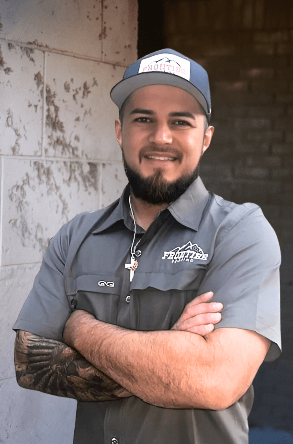 A man with a beard and tattoos wearing a gray shirt and a cap with the logo 'Frontier' standing with arms crossed in front of a brick wall.